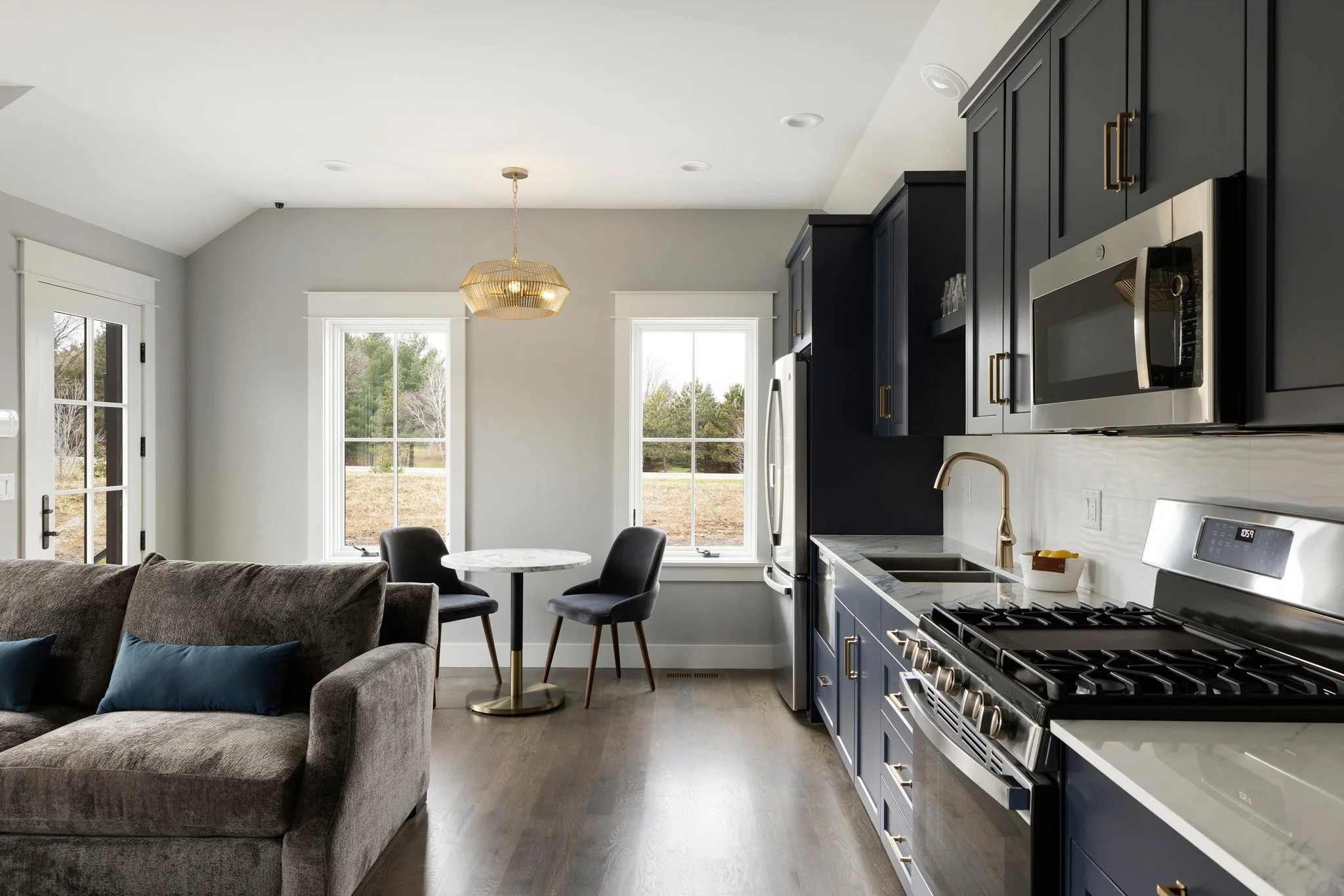 Modern kitchen with navy blue cabinets, stainless steel appliances, a marble countertop, and a living area with a gray couch and a small dining table with two black chairs near large windows.