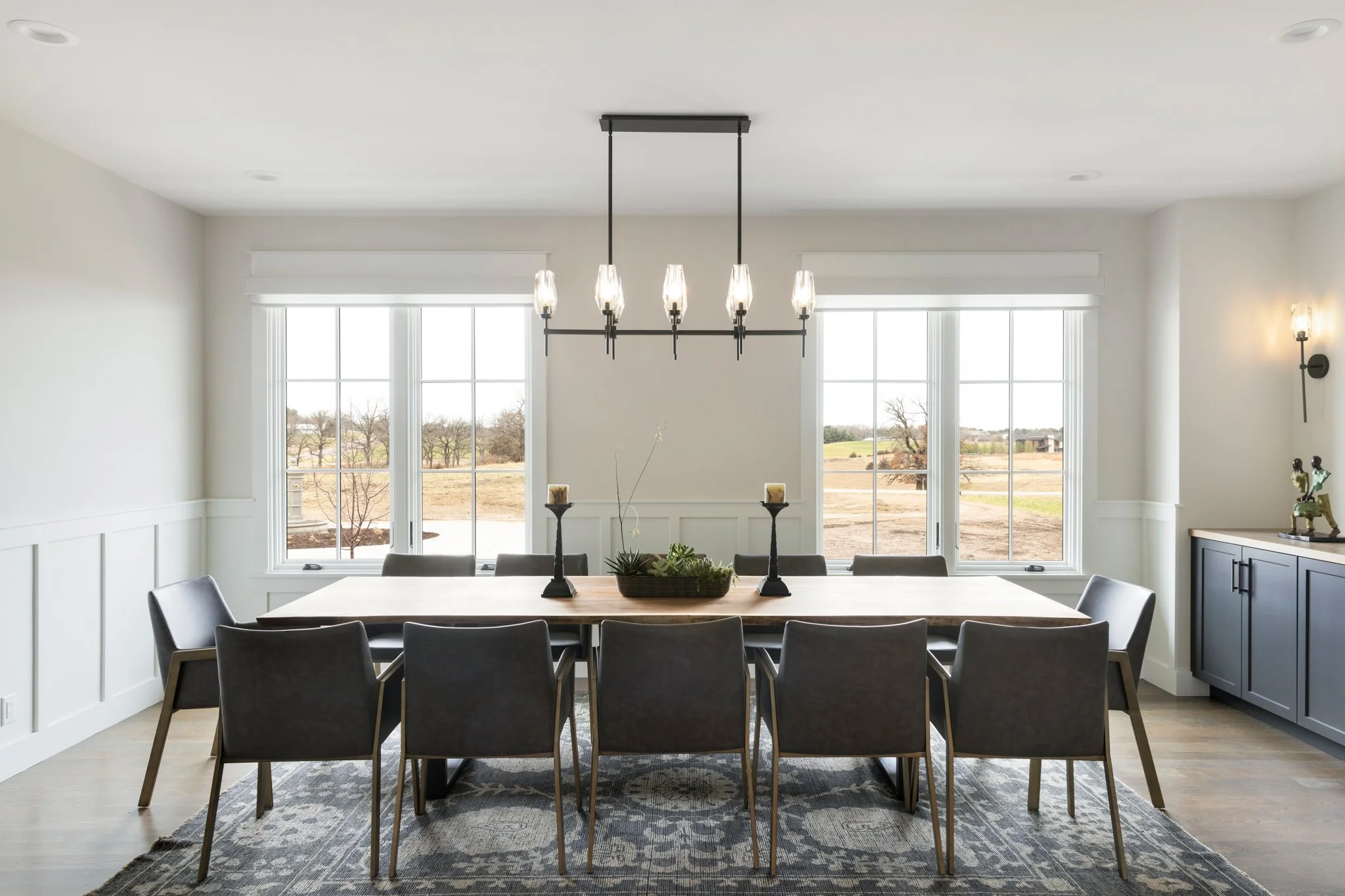 Modern dining room with a large wooden table, nine gray chairs, large windows, a chandelier, and decorative items including candles and a plant.