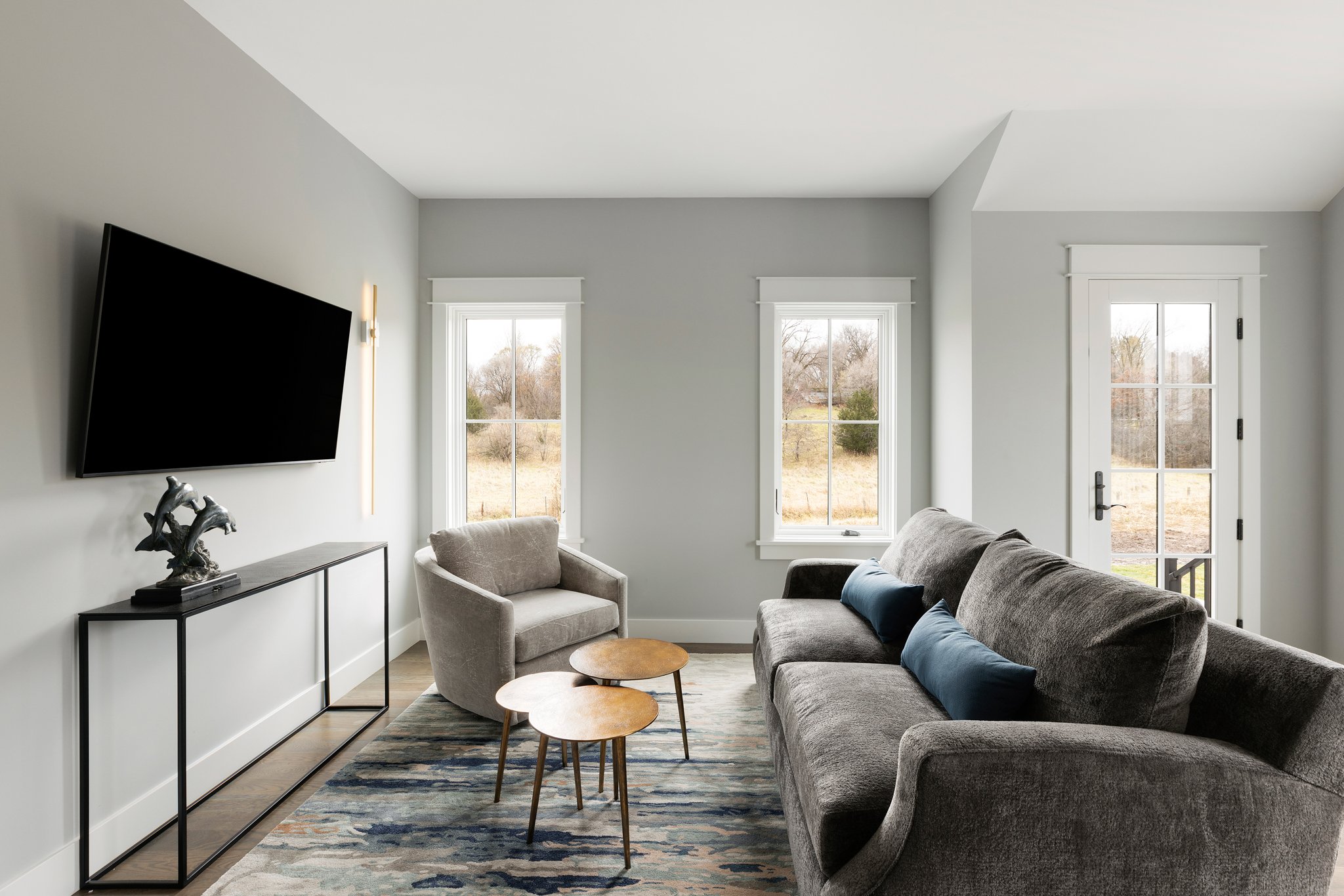 Living room with gray sofa, armchair, two small wooden tables, tv on wall, large windows, door, and view of outdoors.
