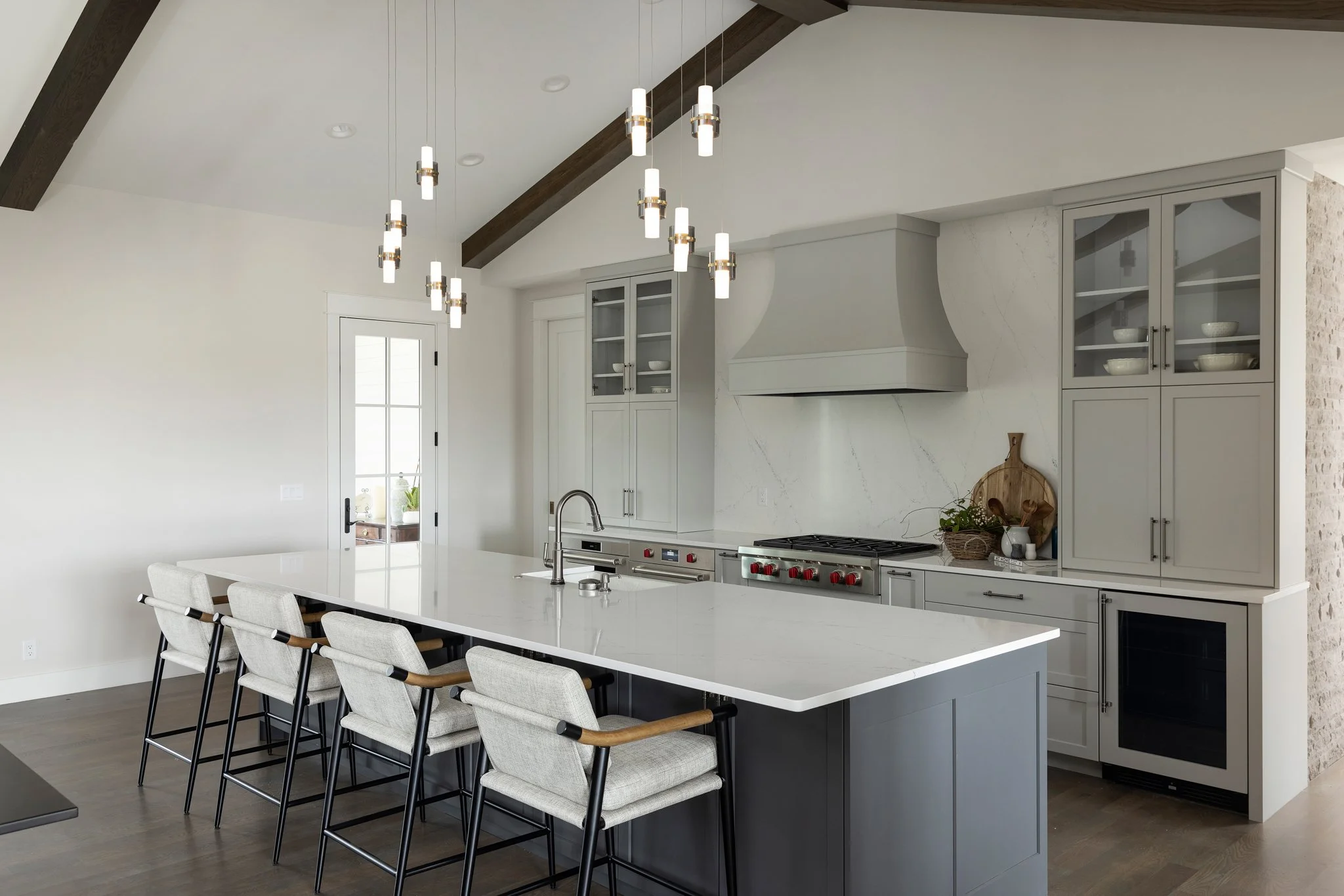 Modern kitchen with white walls, light gray cabinetry, a large white island with a sink, barstools, dark wood ceiling beams, and a hanging light fixture.