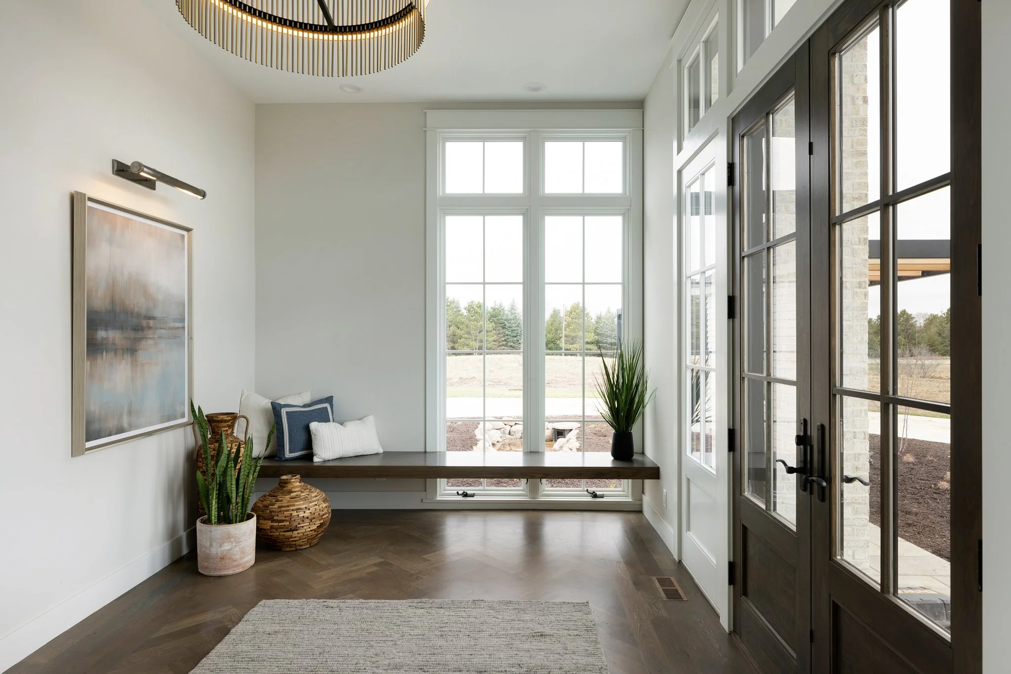Bright sunroom with large windows, wooden bench with pillows, potted plants, and a modern chandelier.