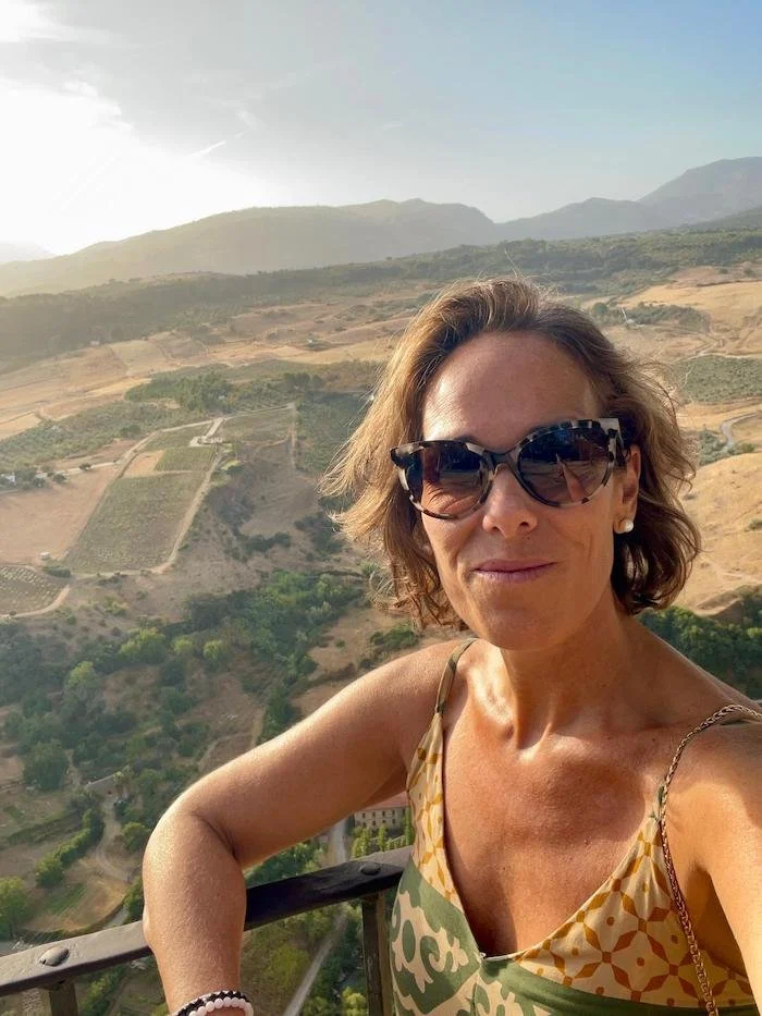 Mujer tomando una selfie en un mirador con campos y colinas en el fondo, durante el día, usando gafas de sol y vestido de verano.