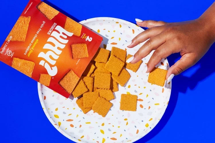 A hand with long white nails reaching towards a white plate filled with square-shaped animal crackers. The crackers are spilling from a red bag labeled 'Why' which is partially visible, on a vibrant blue surface.