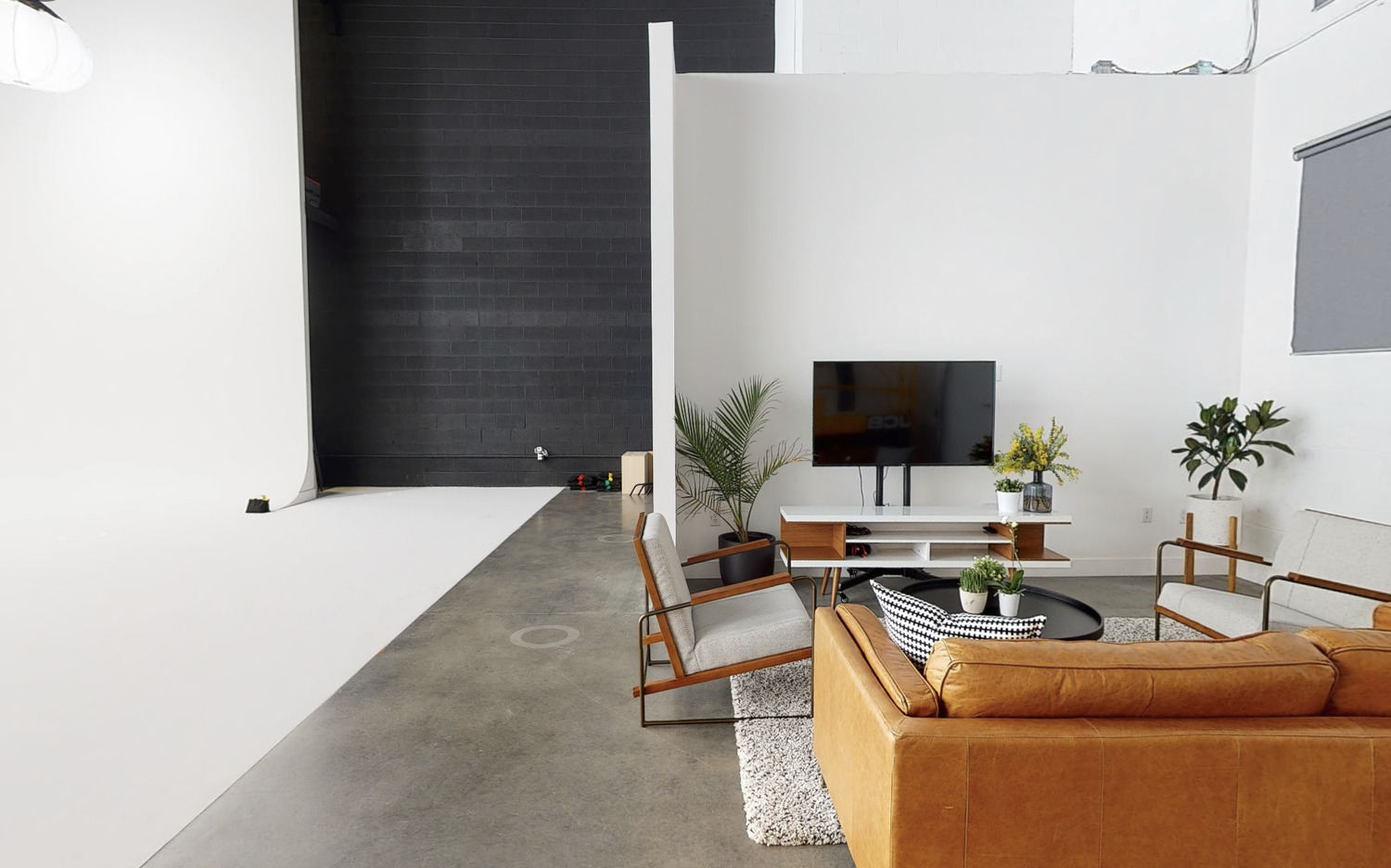 Modern living room with white walls, concrete floor, brown leather sofa, gray armchair, potted plants, wall-mounted TV on a white stand, and large window with gray shade.