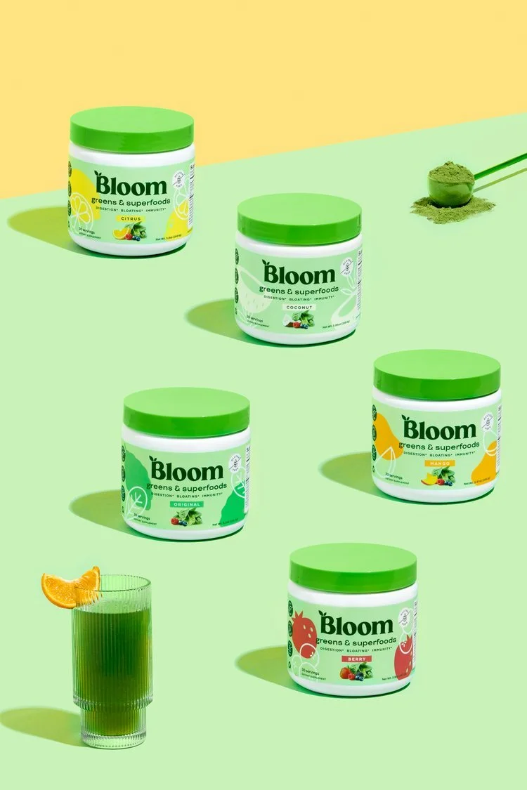 Several containers of Bloom greens and superfoods powder in different flavors on a pastel green and yellow background, with a glass of green juice garnished with an orange slice.