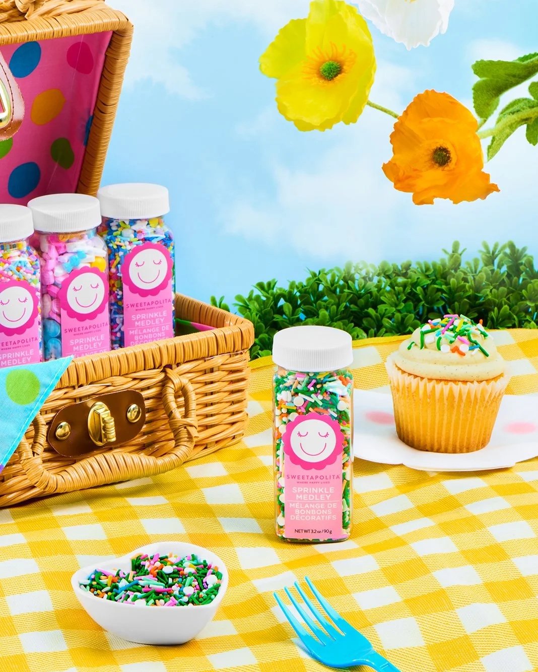 Colorful sprinkles and a cupcake on a yellow checkered tablecloth, with a picnic basket containing candy bottles, a bowl of sprinkles, and a fork, outdoors with flowers and greenery in the background.