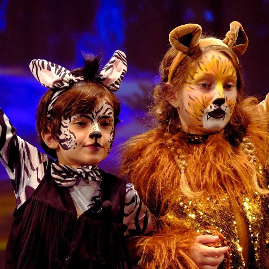 Four children dressed in costumes from 'The Wizard of Oz' perform on stage with a colorful countryside backdrop.