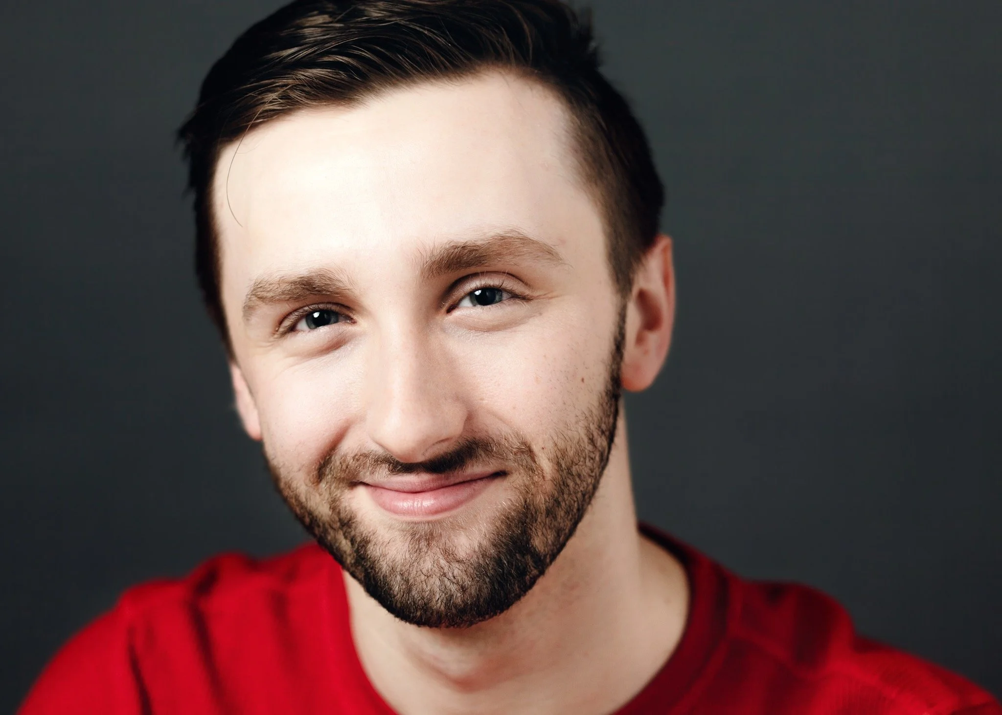 Close-up of a smiling young man with dark hair and a beard wearing a red shirt against a dark background.