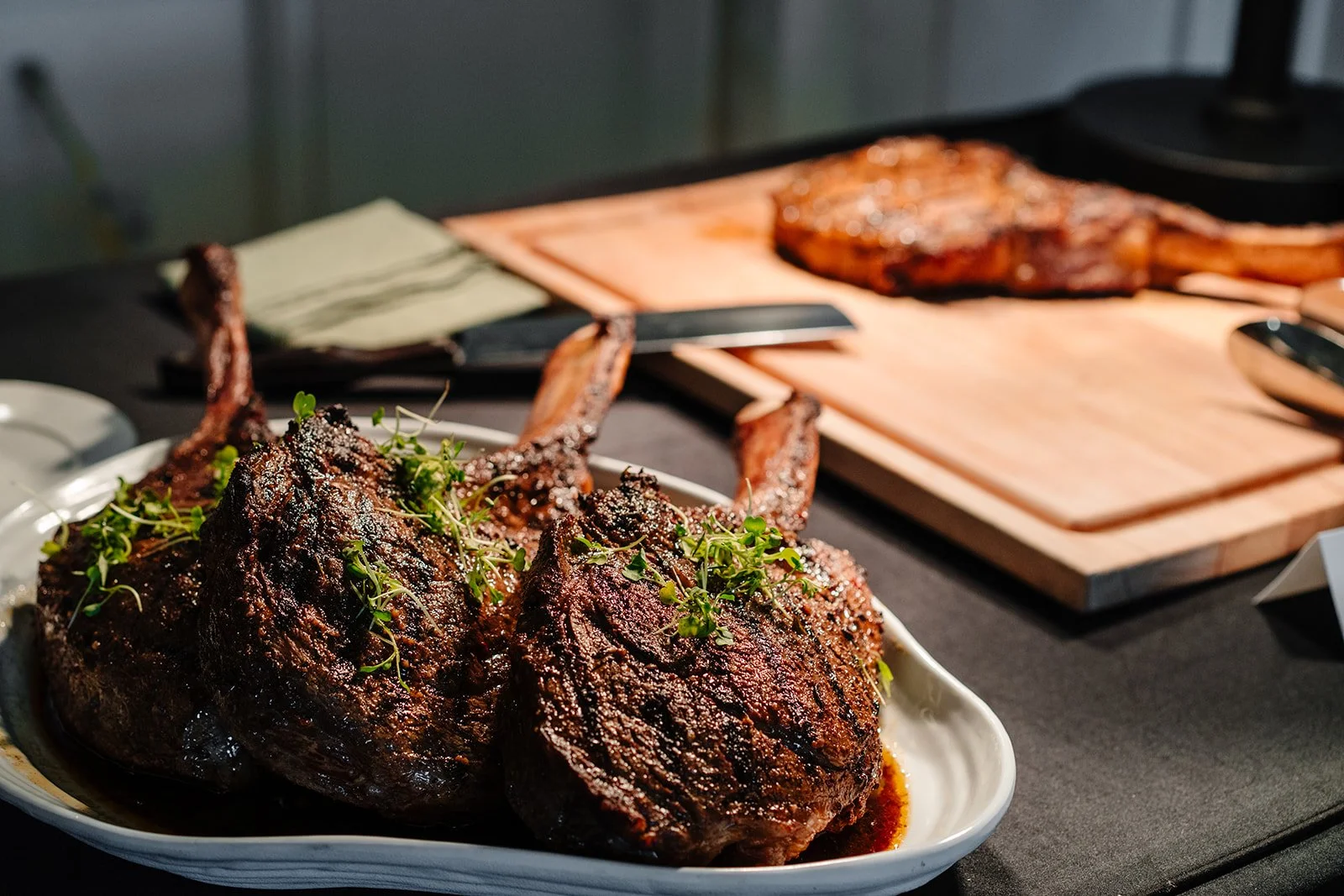 Cooked lamb chops garnished with greens on a white plate, with a wooden cutting board and more meat in the background.