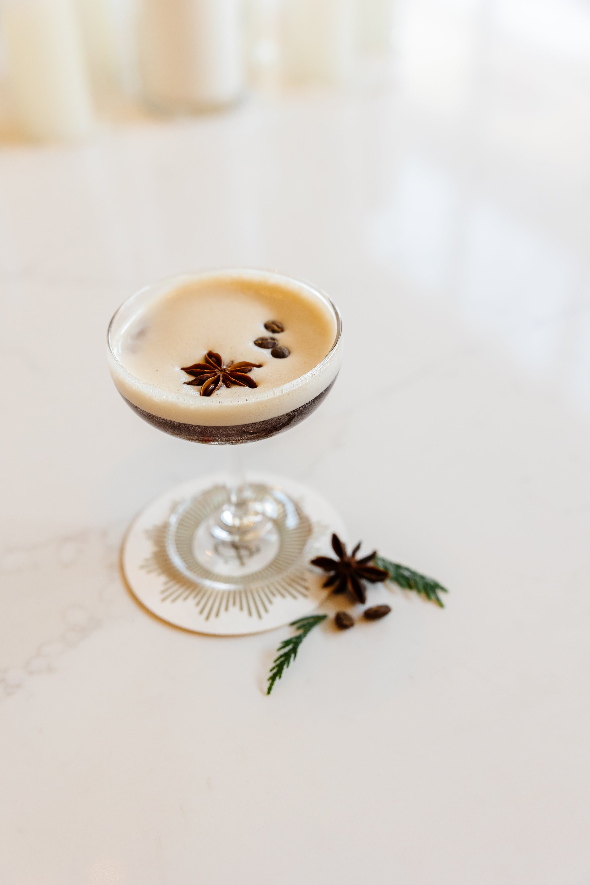 A cocktail glass with a frothy drink garnished with star anise, coffee beans, and a sprig of greenery, placed on a white surface with a decorative coaster.