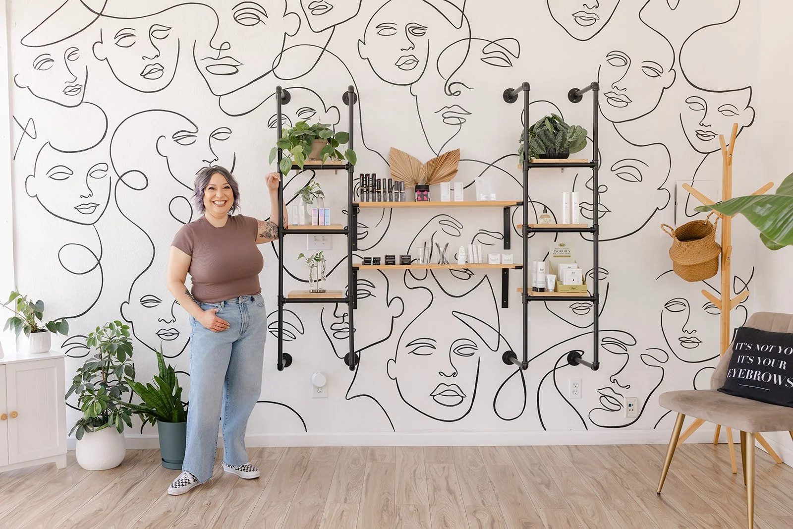 A woman with short gray hair, wearing a brown t-shirt and light blue jeans, smiling and standing in a decorated room. The room has a wall with a black line art mural of abstract faces, wooden and metal shelving with plants and skincare products, a po