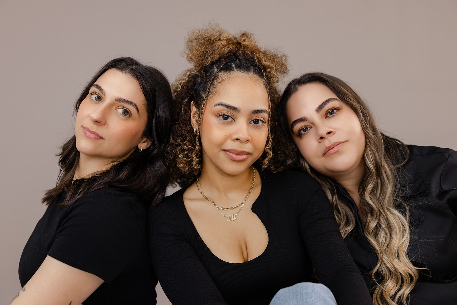 Three women sitting close together against a neutral background, facing the camera.