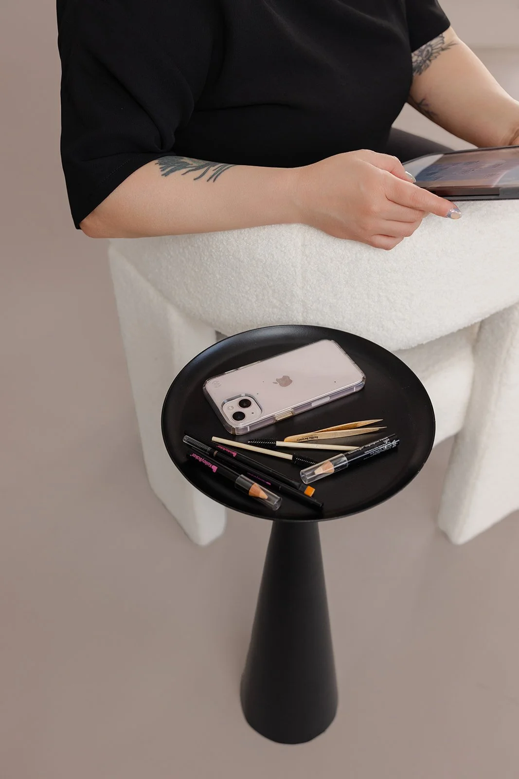 A person with tattoos on their arm is sitting on a white chair, using a tablet. A black round side table in front holds an iPhone, pens, brushes, and makeup tools.