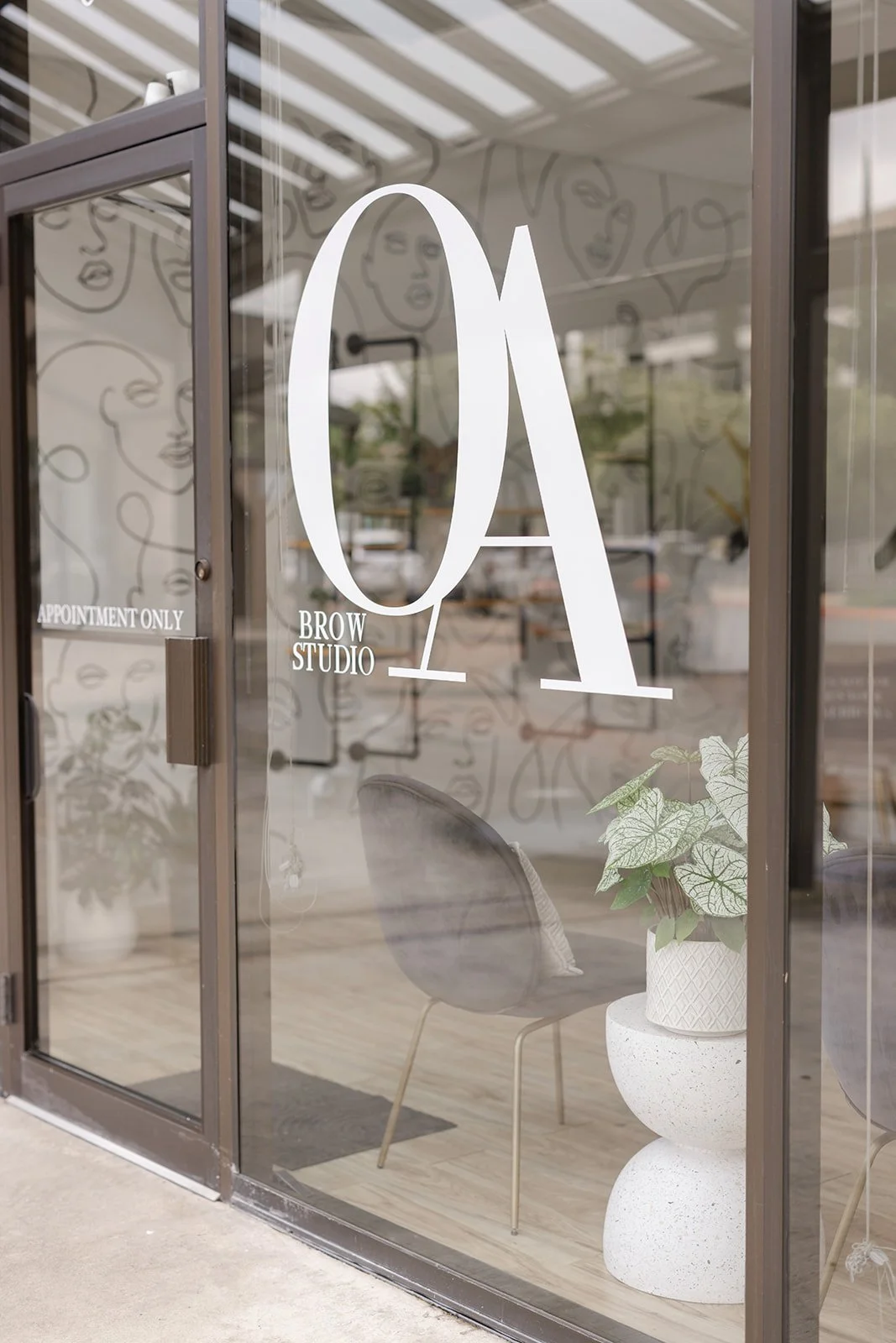 Glass door with the logo 'QA Brow Studio' and a small sign that says 'Appointment Only', a chair, and a potted plant inside.