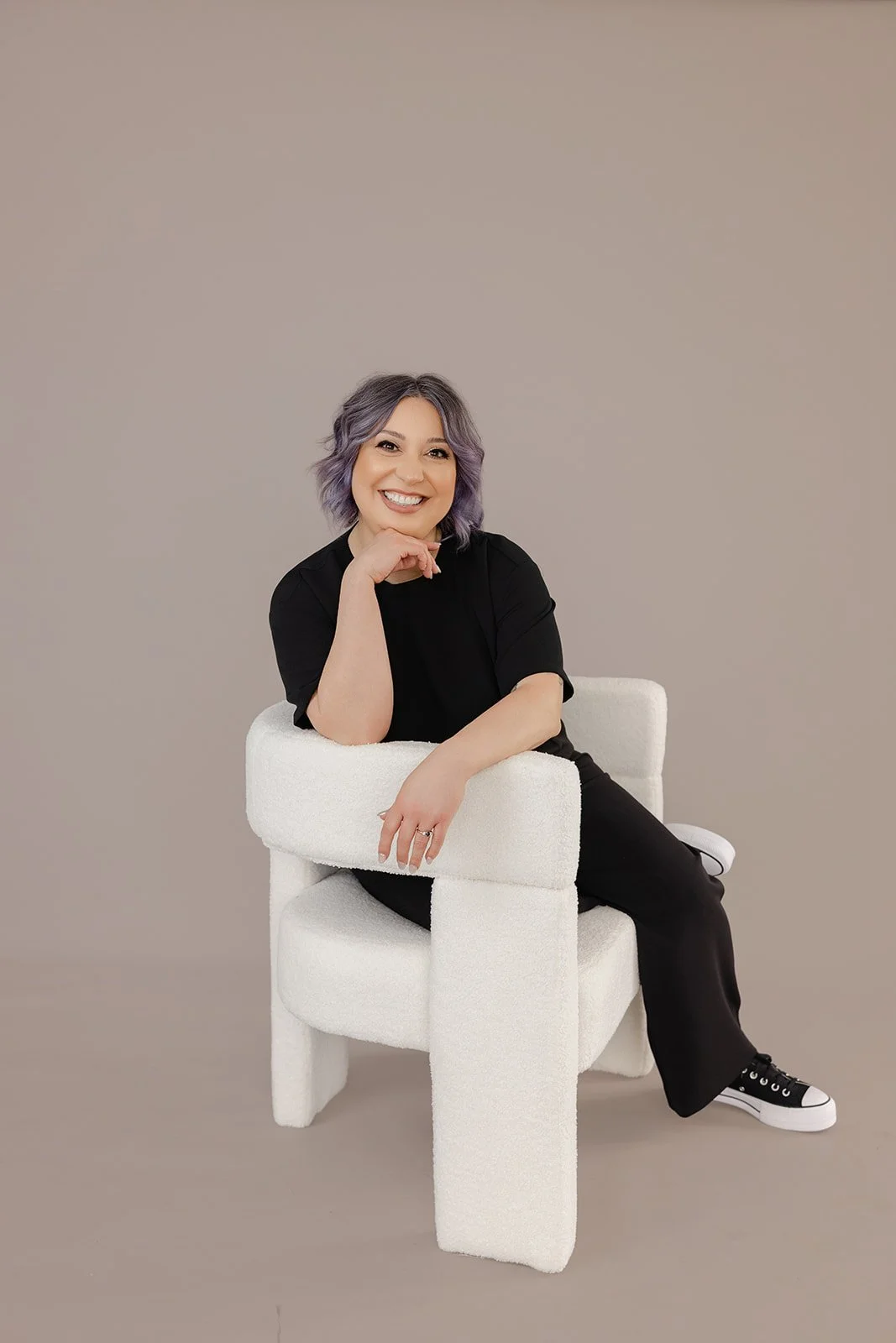 A woman with shoulder-length purple hair, wearing a black shirt and black pants, sitting on a white modern chair with a beige background.