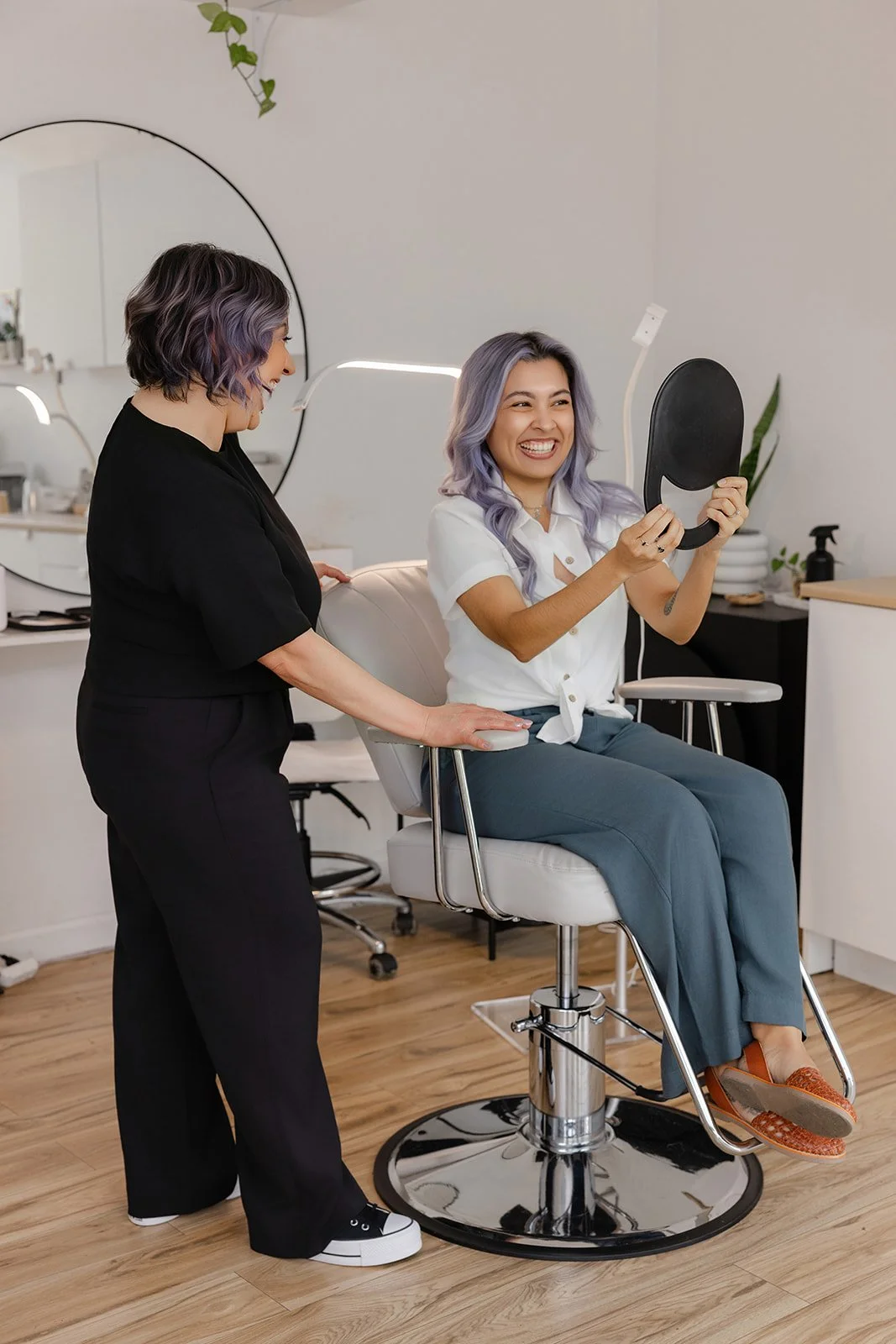A woman with purple hair sitting in a salon chair, holding a mirror and smiling, with a stylist standing beside her in a black outfit.