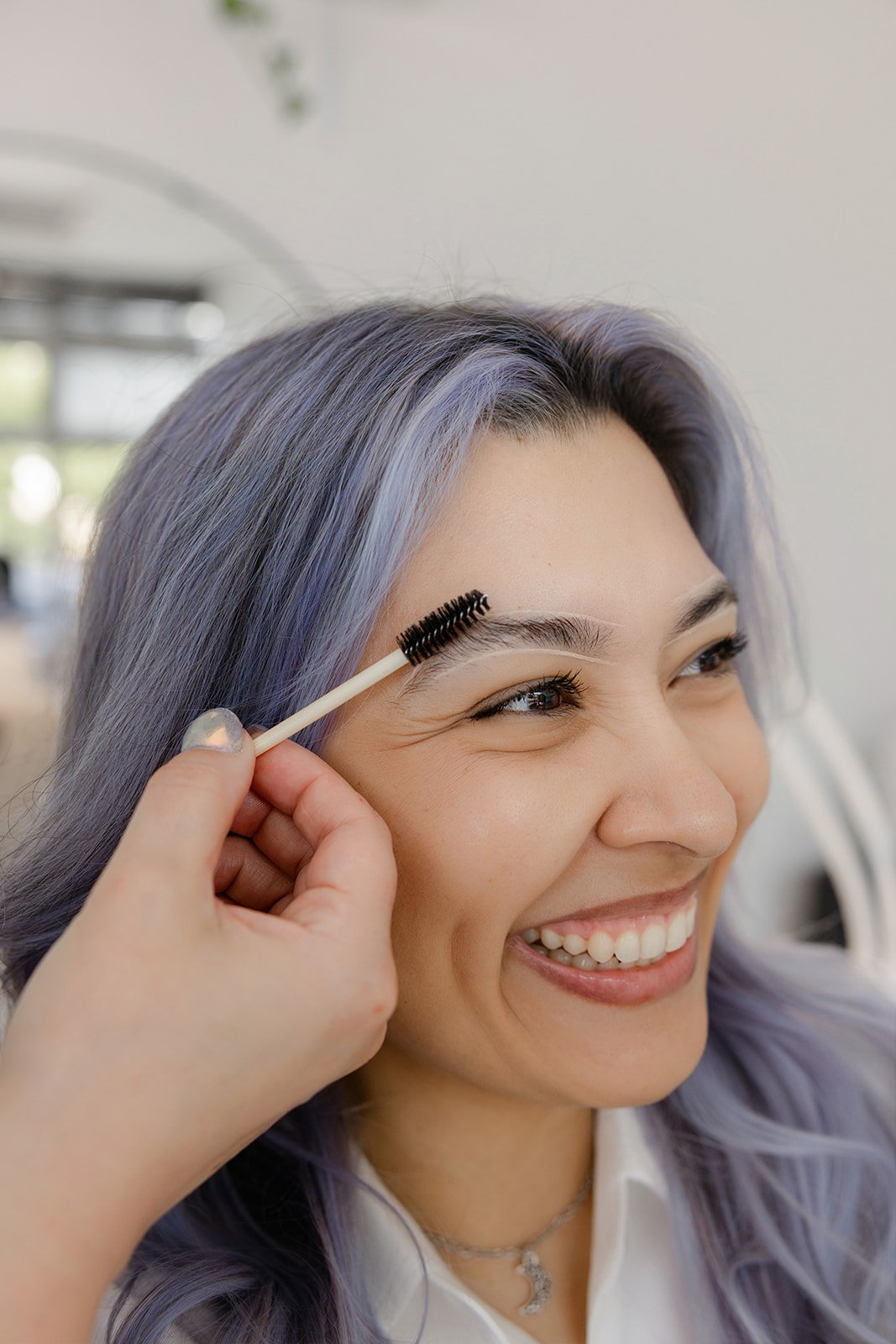 Woman with grayish purple hair getting eyelashes curled while smiling.