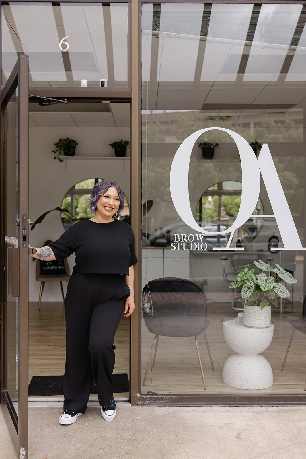 A woman with shoulder-length purple hair, wearing a black top and black pants, stands in the doorway of a salon called Oa Brow Studio, smiling. There are potted plants and chairs inside the salon, visible through the glass window.