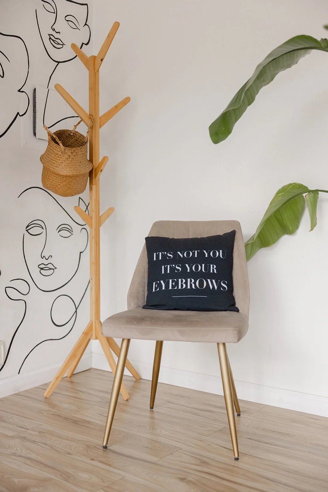 A beige chair with a black pillow that says, 'It's not you, it's your eyebrows,' a wooden coat rack with a woven basket hanging from one of its hooks, a minimalist line art wall drawing of abstract faces, and a large green plant with broad leaves in 