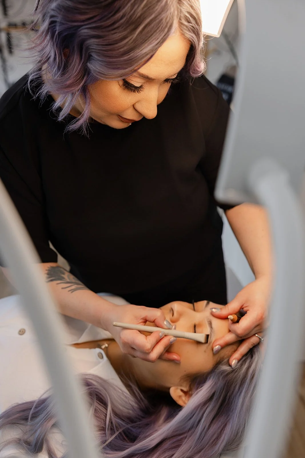 A woman with purple hair doing makeup or cosmetic procedure on a client's face in a professional setting.