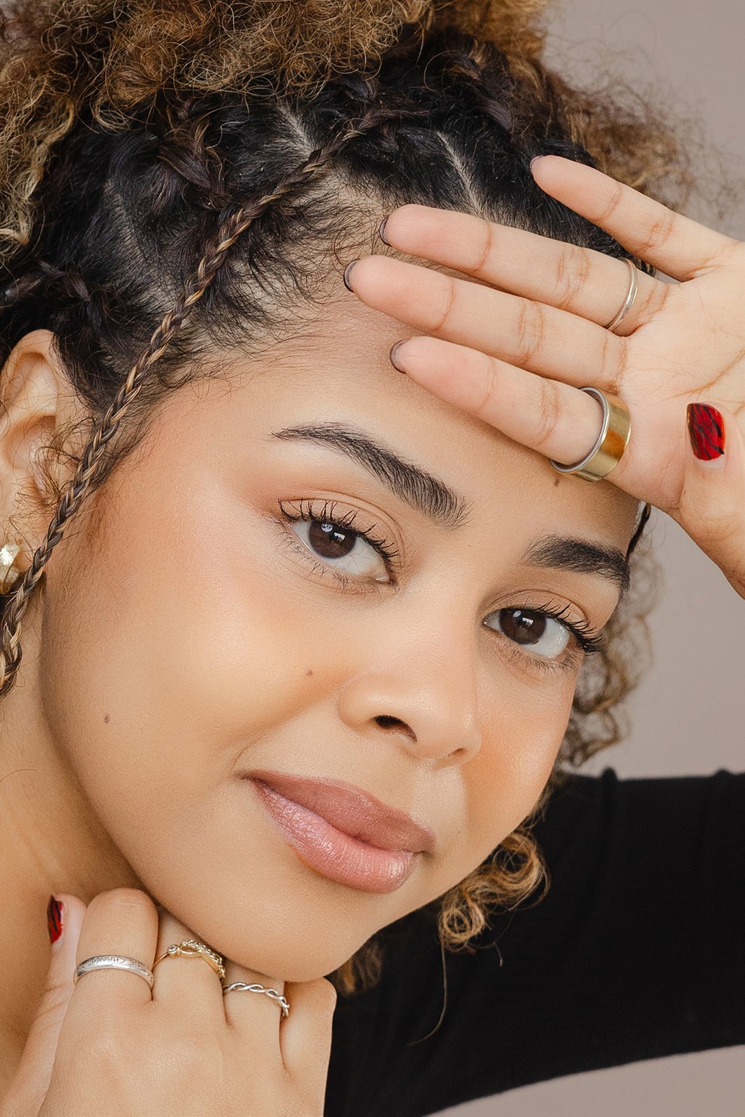 A woman with curly hair and brown eyes posing with her hand on her forehead showing off her eyebrows, wearing rings and nail polish, smiling softly.