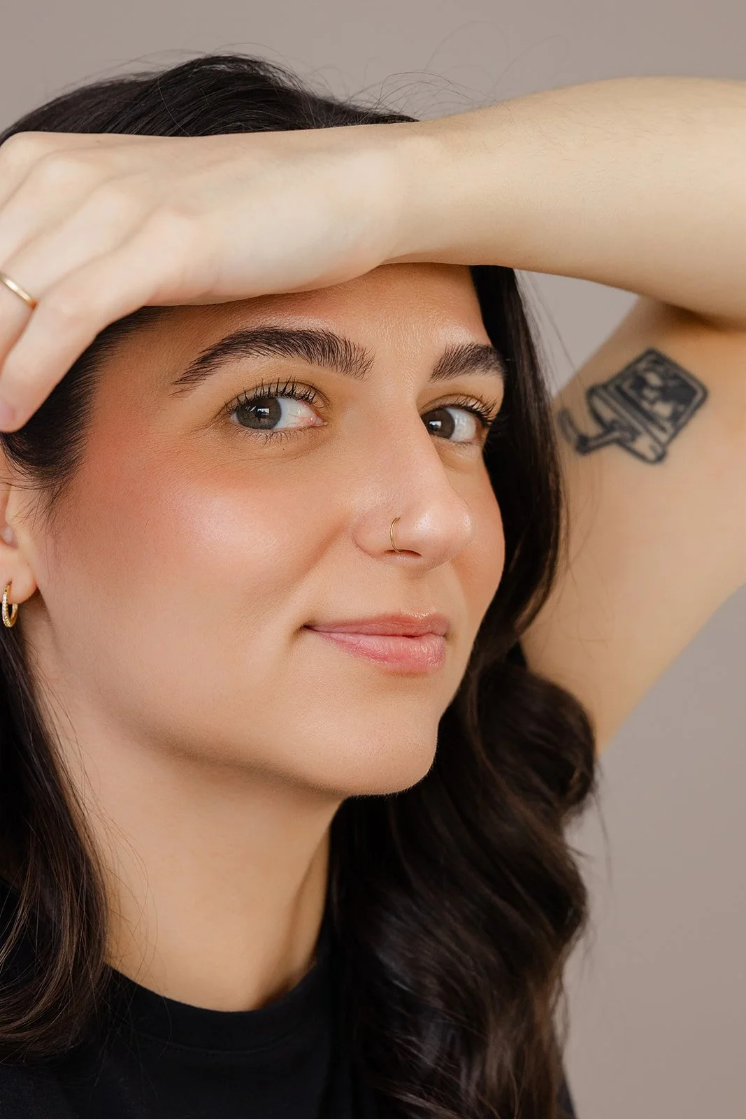 Close-up of a woman with dark hair and blue eyes, wearing a nose ring and gold earrings, with one arm raised and hand resting on her forehead, showing a tattoo on her upper arm.