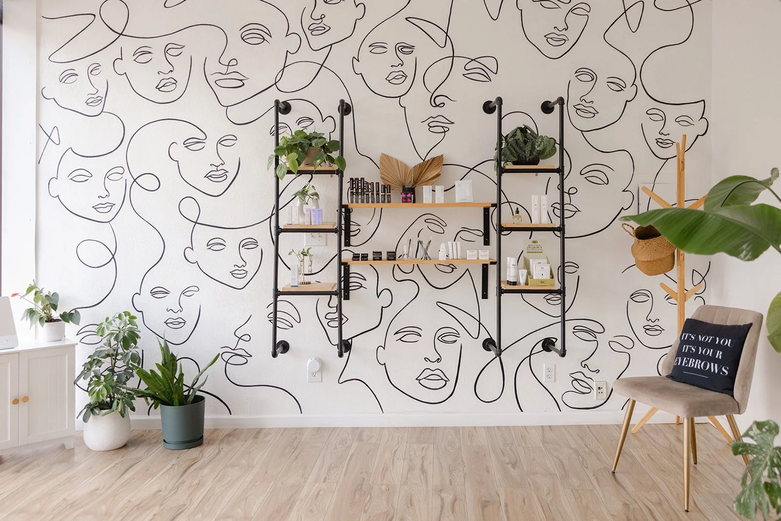 Interior of a minimalist boutique with a wall decorated by continuous black line art drawings of women's faces. There are wooden and black metal shelves with plants and beauty products, a beige chair with a black cushion that has the quote "It's not 