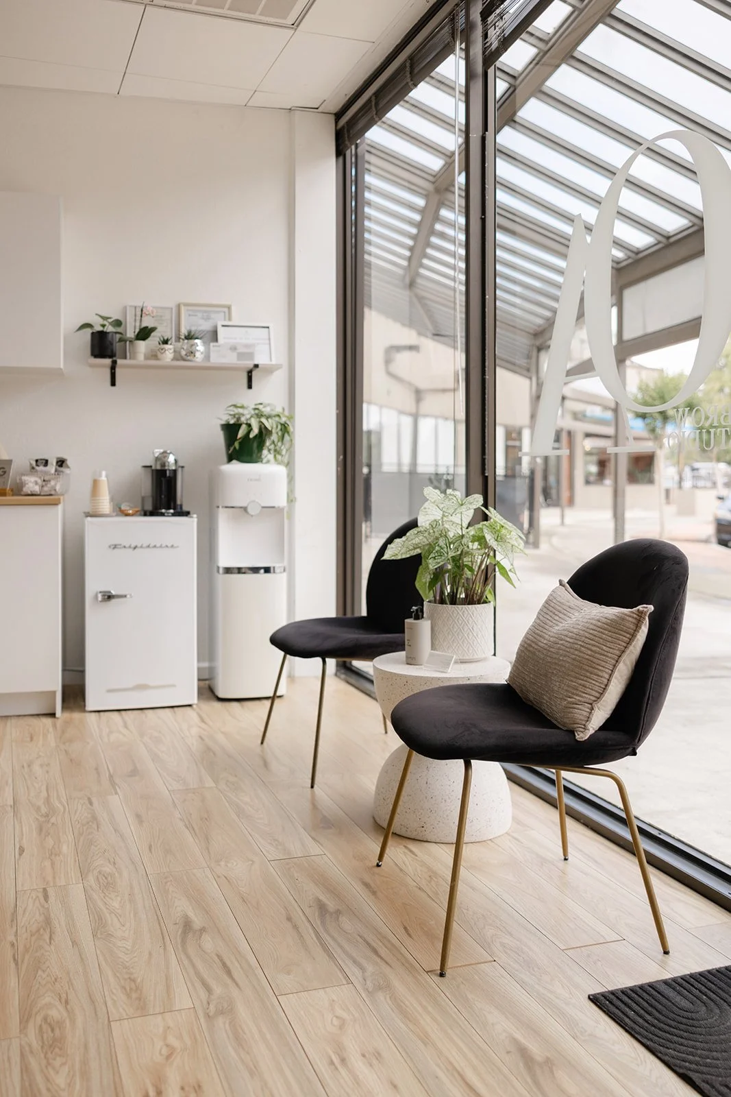 Interior of a modern coffee shop with black chairs, a small side table with a plant, a water dispenser, and large glass windows.