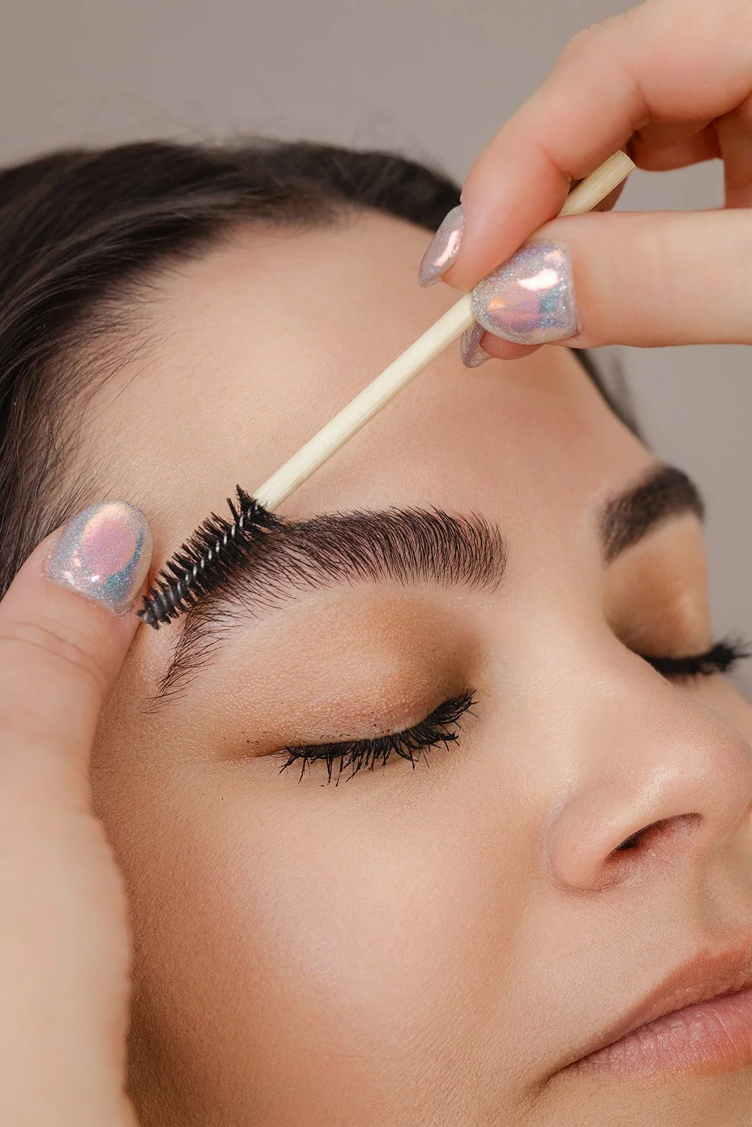 Close-up of a woman with closed eyes getting her eyebrows groomed with a spoolie brush by a person with glittery nail polish.