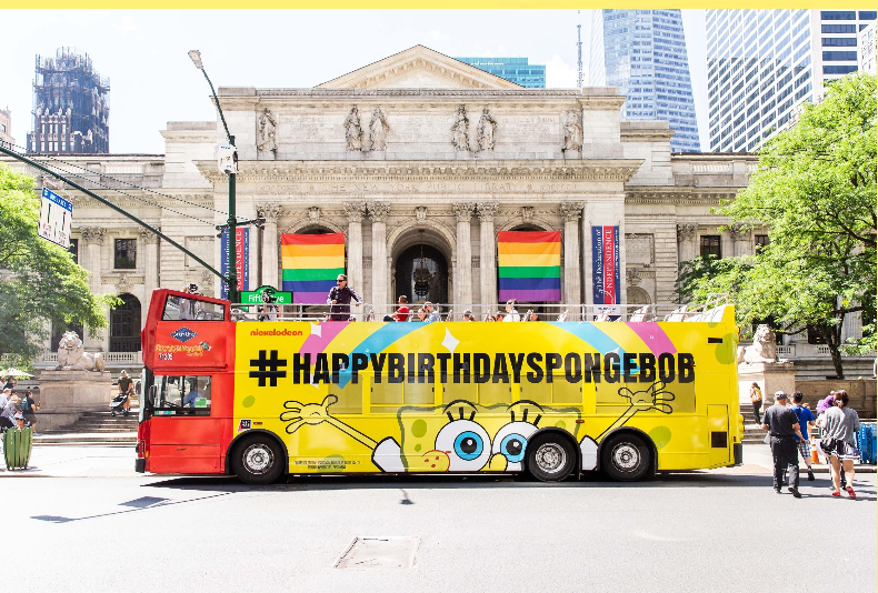 A yellow Spongebob-themed birthday parade float with the hashtag #HAPPYBIRTHDAYSPONGEBOB parked in front of a historic building with rainbow flags, in an urban city setting with trees and tall buildings.