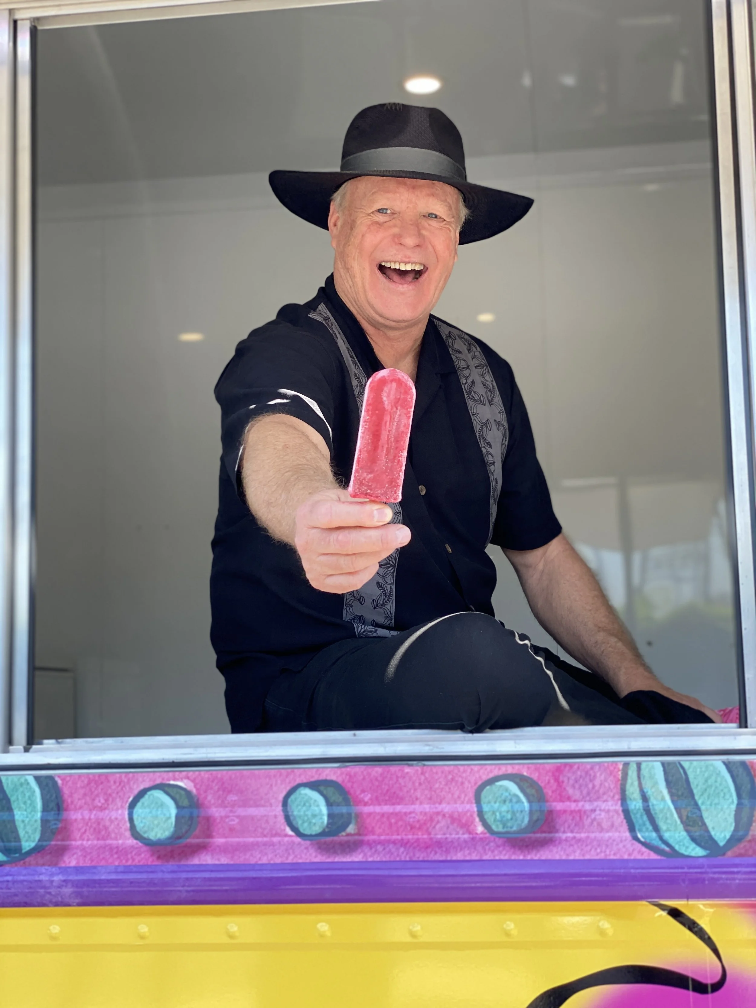 A man wearing a black hat and black shirt sitting in an ice cream truck, smiling and holding a pink popsicle towards the camera.