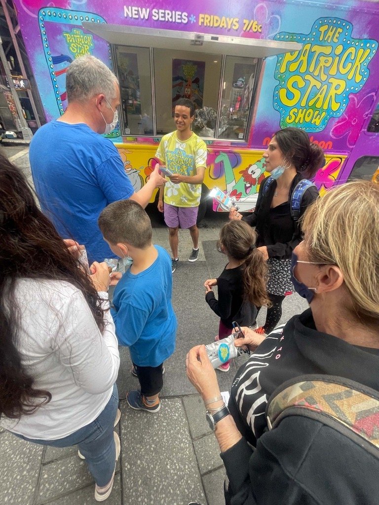 A group of people gathered around a man in front of a colorful trailer for 'The Patrick Star Show' on a city street. The man is smiling and handing something to the man in a blue shirt, while the others look on. Some people are wearing masks.
