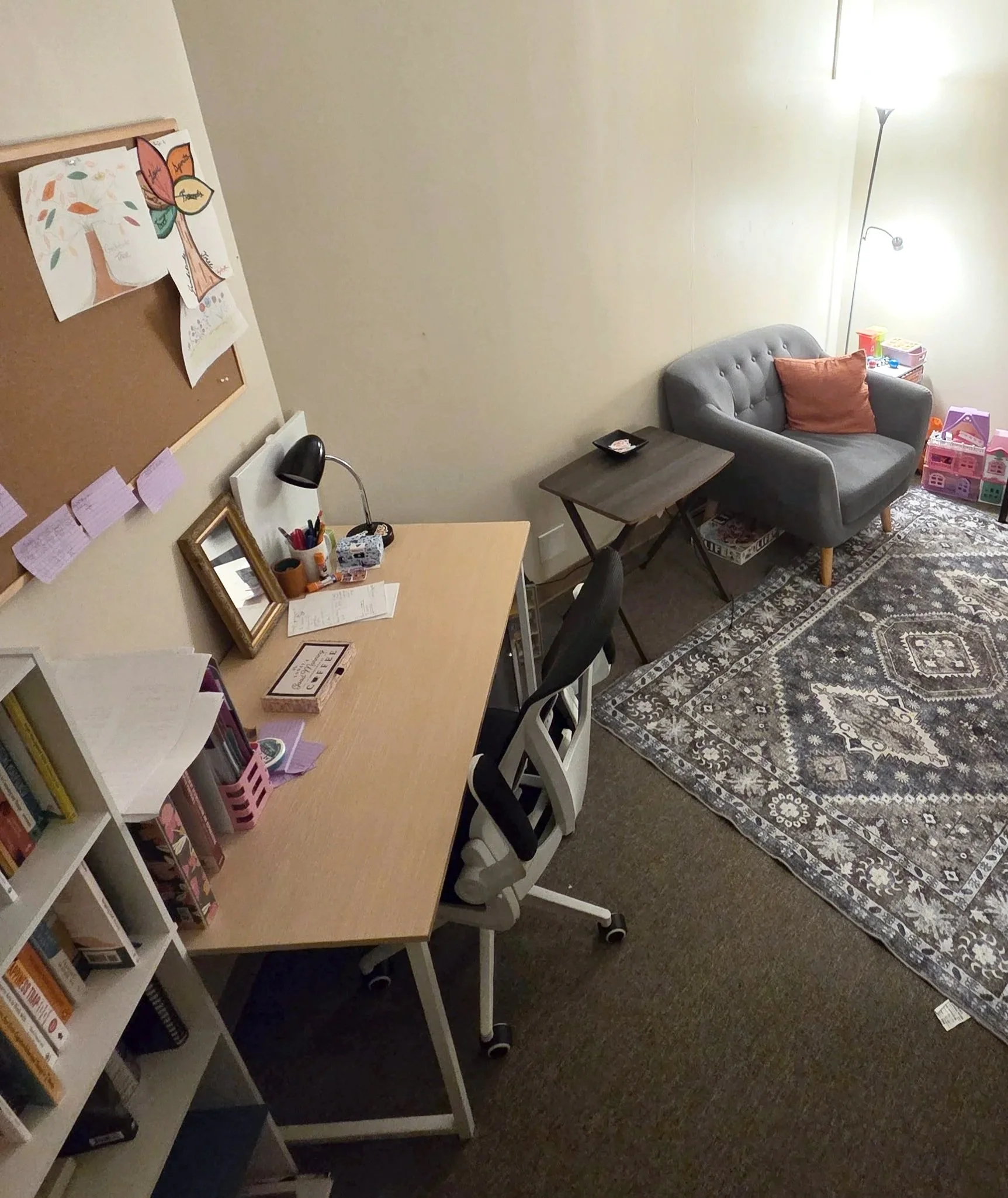 View of a cozy office area with a wooden desk, office chair, cork board with notes, a gray loveseat with orange pillow, small side tables, and toys, illuminated by a tall floor lamp.