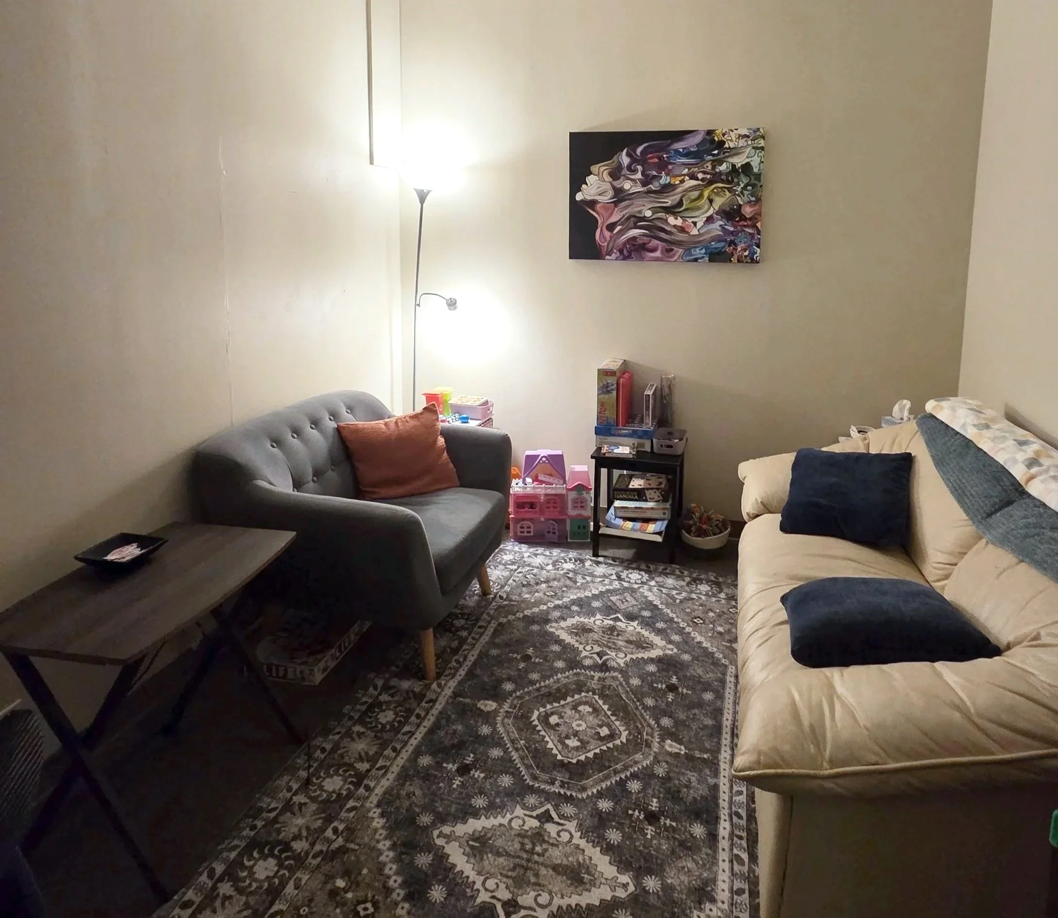 A cozy office space with a beige sofa and a gray loveseat, a patterned area rug, a small black side table.  A tall floor lamp provides lighting, and there's children's toys and books on a black shelf in the corner.