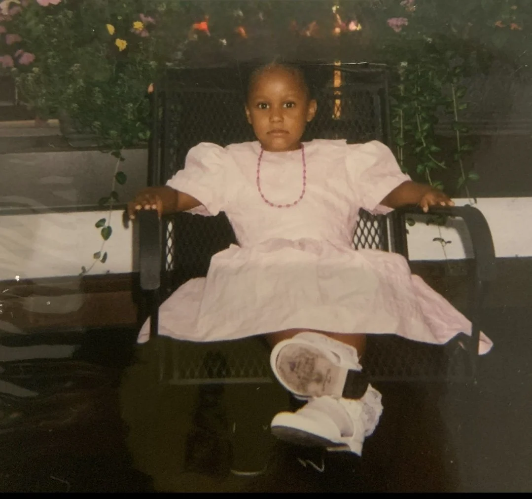 A young girl in a pink dress sitting on a black outdoor bench with her arms resting on the armrests, looking directly at the camera. She is wearing a beaded necklace and matching shoes, and there are plants and flowers in the background.