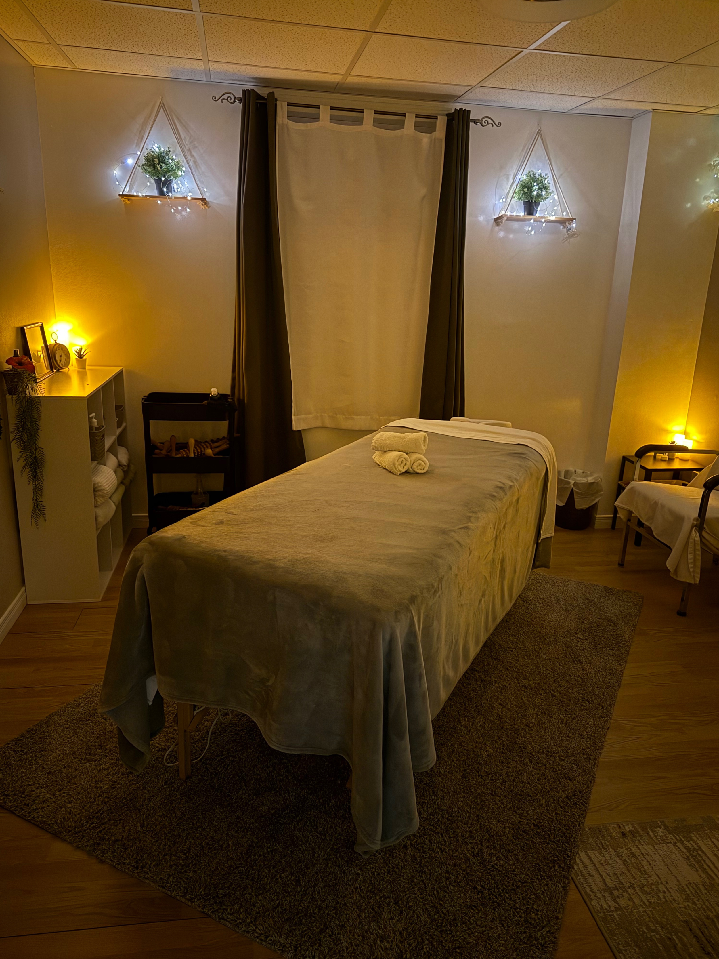 Salle de massage avec lit recouvert d'une couverture beige, serviettes pliées dessus, murs éclairés par deux lampes, décorations végétales, rideaux noirs, et petites étagères avec des plantes et des objets