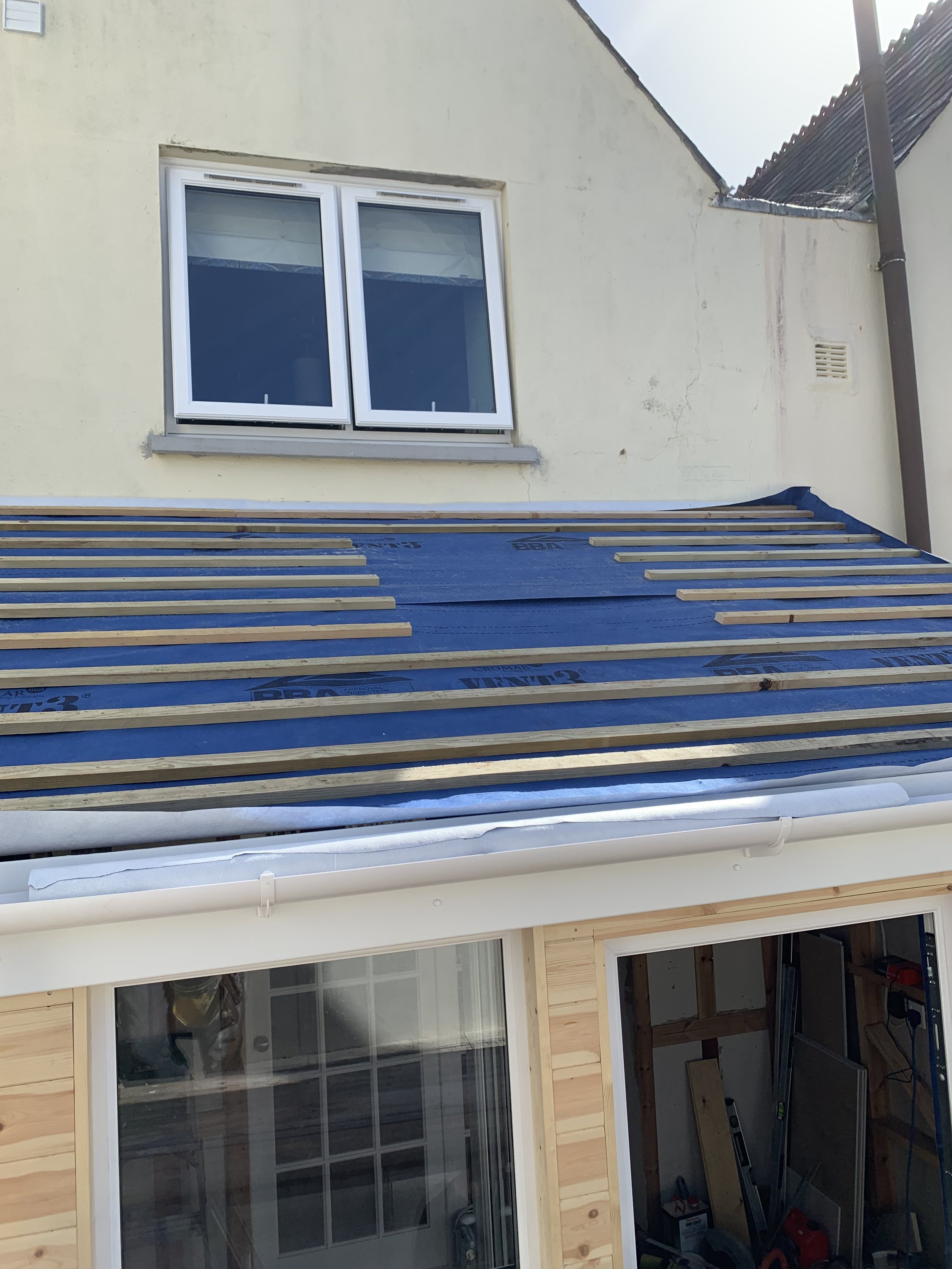A house under renovation showing an upper window, part of the roof with construction materials, and a lower section with a large window and construction tools inside.