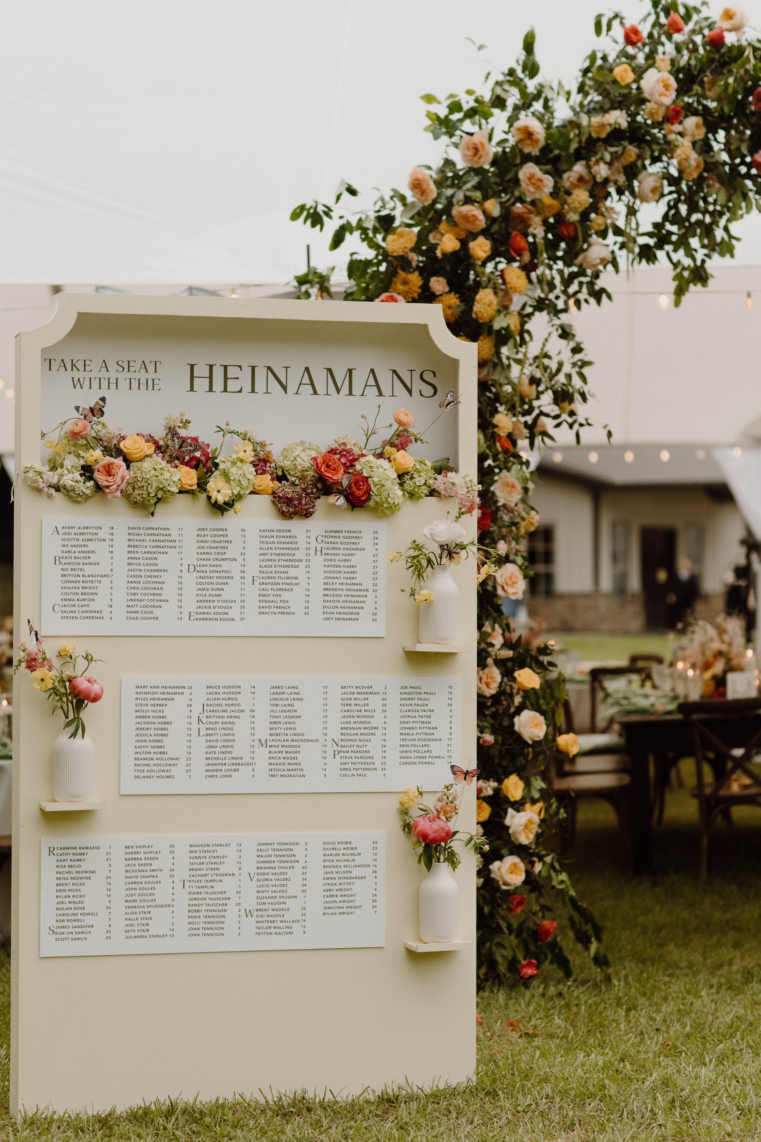 Wedding seating chart decorated with white vases holding flowers, behind a floral arch and surrounded by flowers and string lights - Reception Details