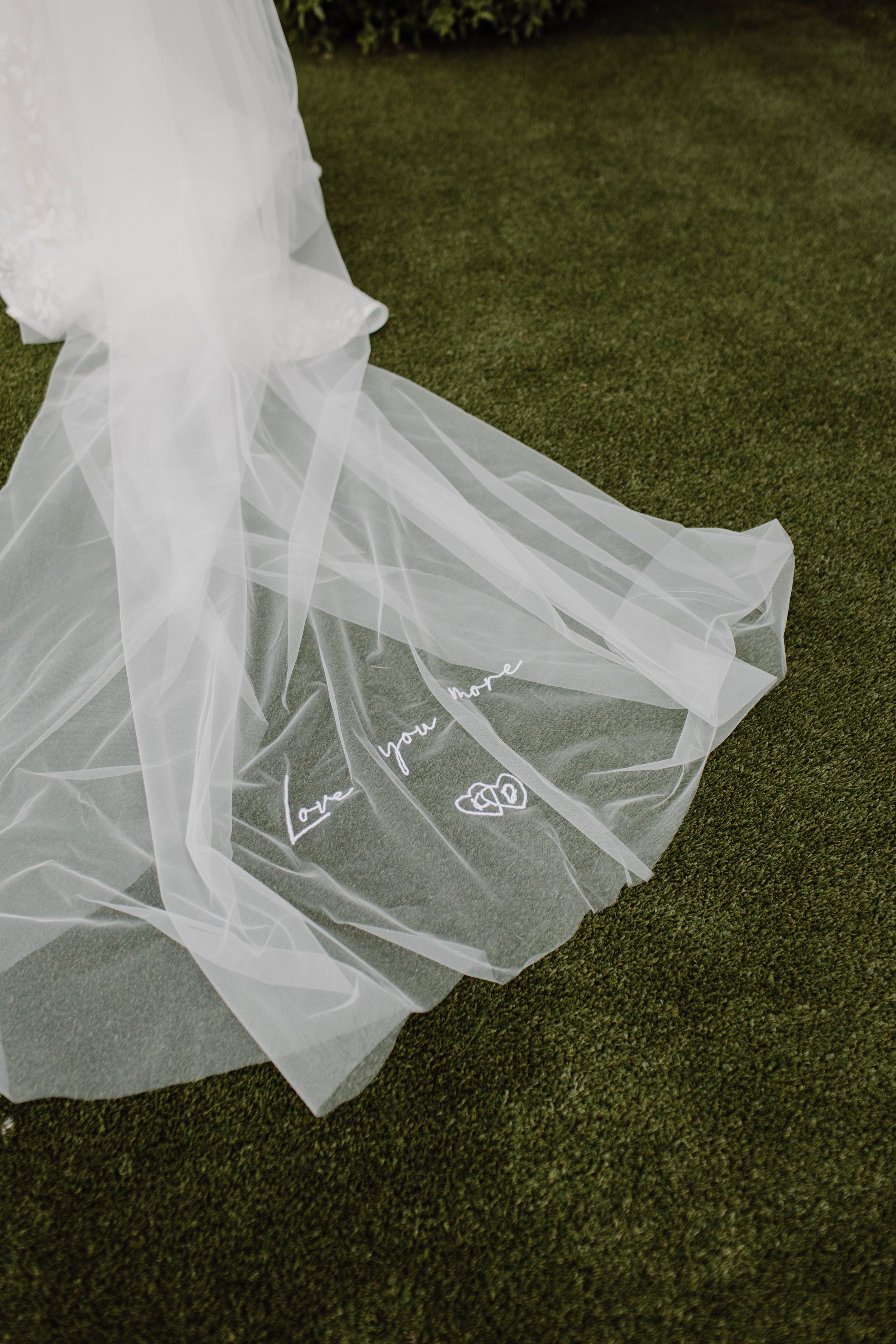 A long, white, tulle veil with the words 'Love you more' and two hearts embroidered on the edge, lying on green grass - showing off the little details that mean so much.