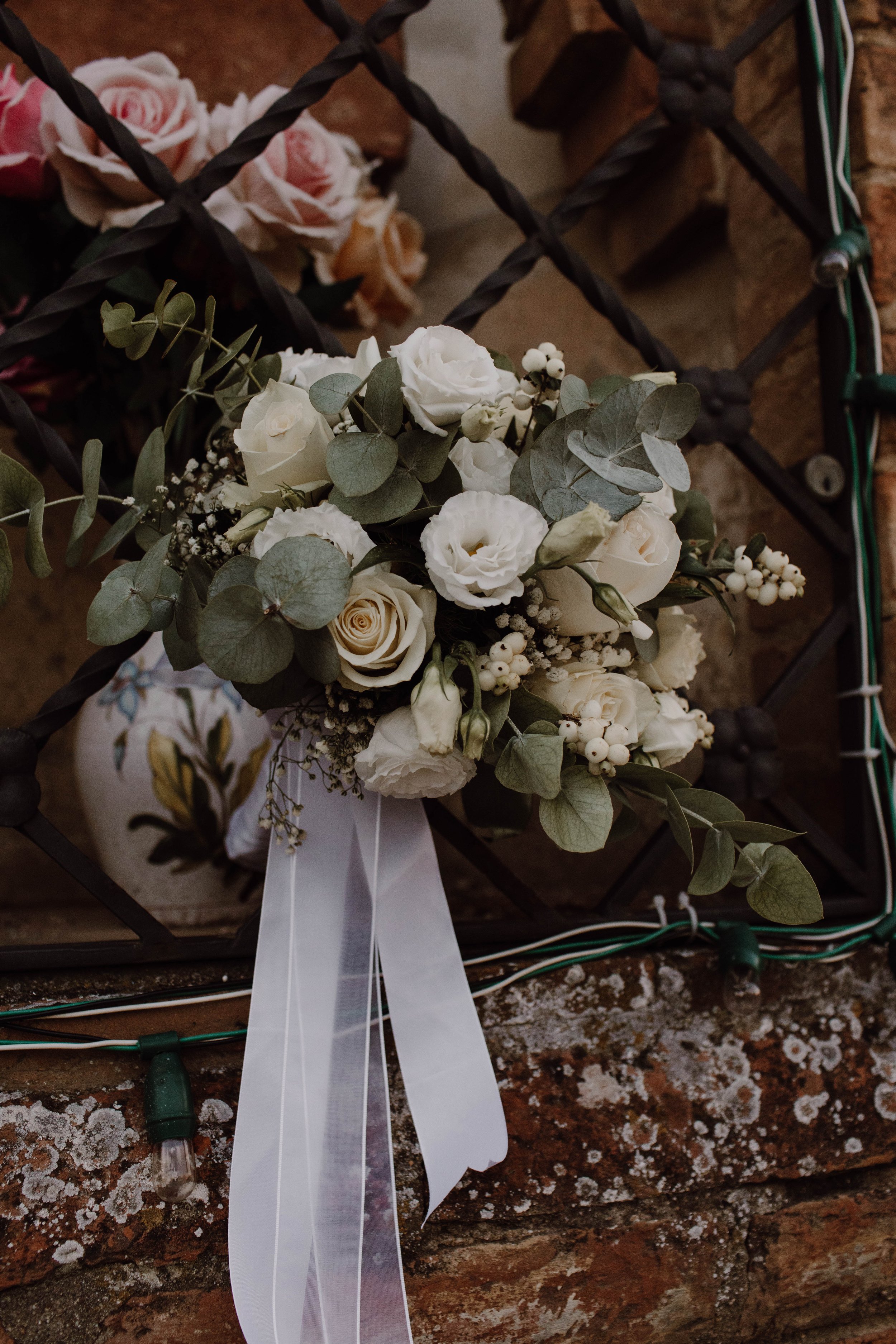 A bouquet of white roses, lisianthus, and greenery with white ribbon, resting on a brick ledge with some branch lights and another floral arrangement in the background.