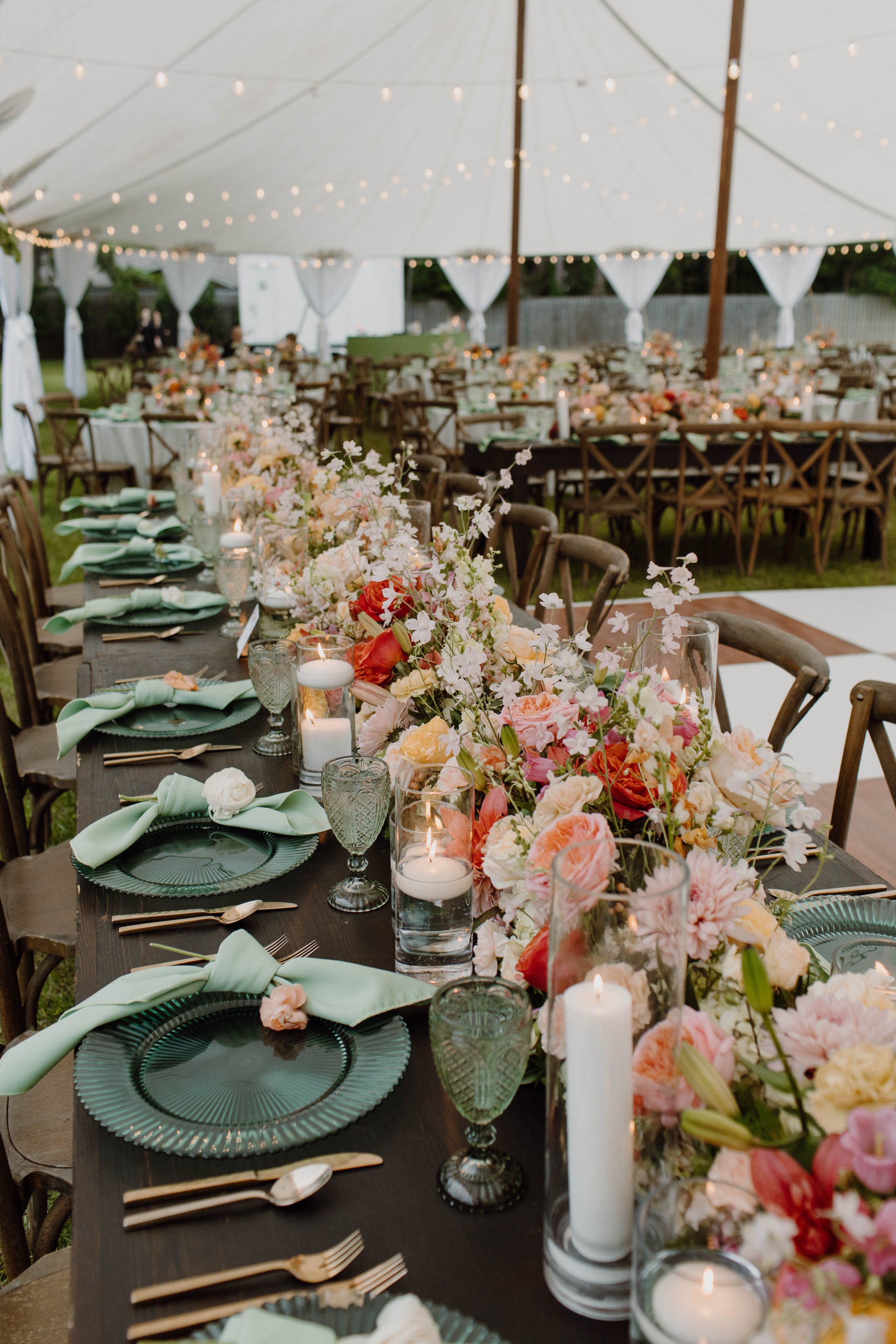 Elegant outdoor event setup with a long table adorned with floral arrangements, candles, and pastel green napkins, under a decorated tent with hanging string lights.