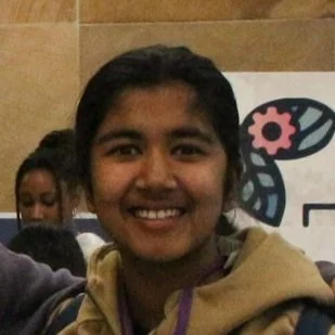 Smiling person with dark hair in an indoor setting, with a crowd and a sign featuring a butterfly logo in the background.