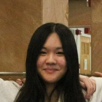 A woman with long black hair smiling indoors, with a wooden wall and some doors in the background.