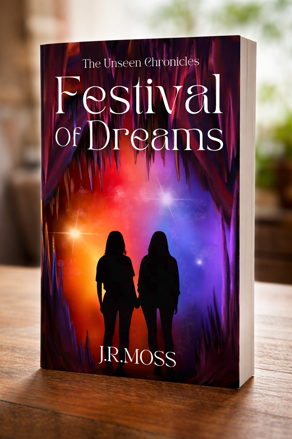 Book titled "Festival of Dreams" from the series "The Unseen Chronicles" by J.R. Moss, featuring silhouettes of two women holding hands against a colorful, dreamy background with sparkling lights and featuring dark, pointed shapes at the top.