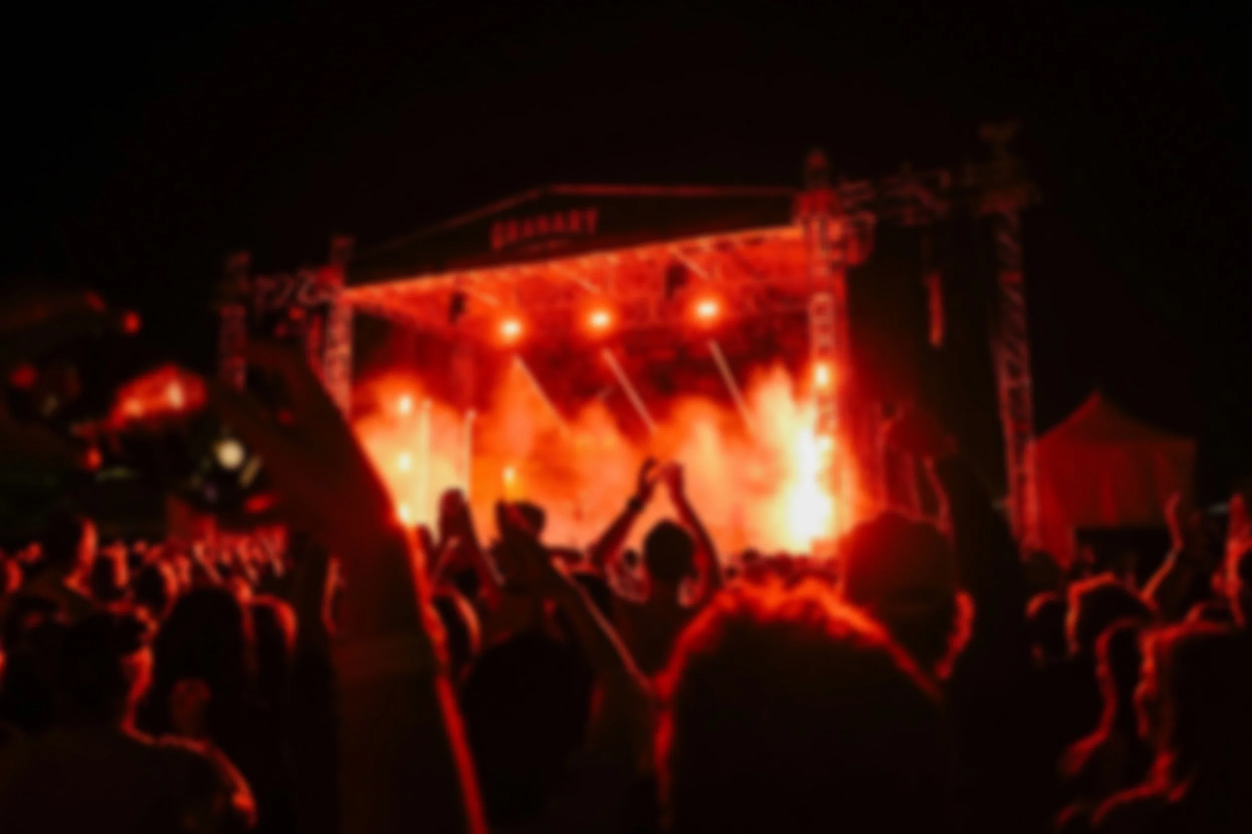 Night scene of a crowd at a music concert with a large stage illuminated in red and orange lights, with smoke and flames visible.