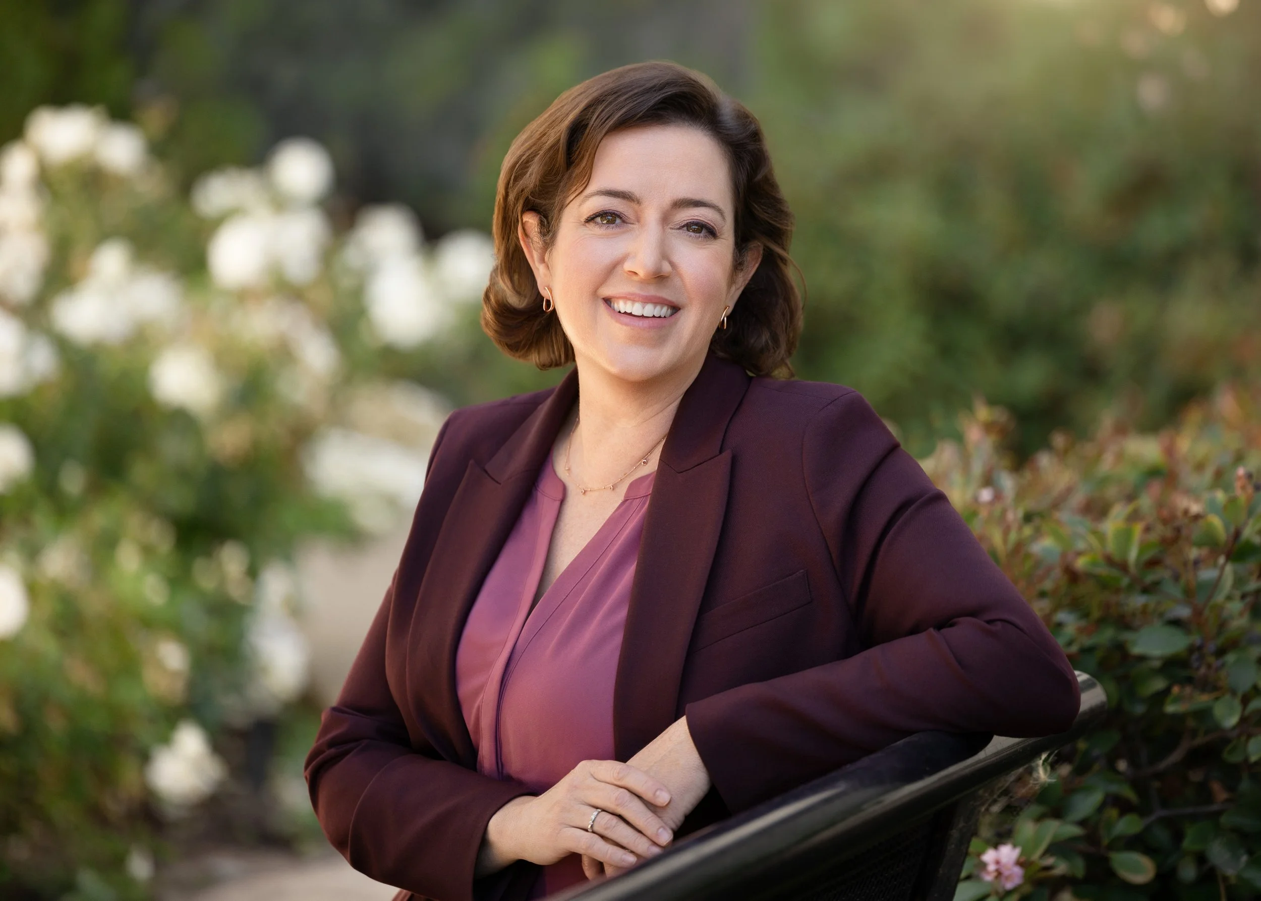 A woman with shoulder-length brown hair, wearing a purple blazer and earrings, smiling outdoors near greenery and flowers.