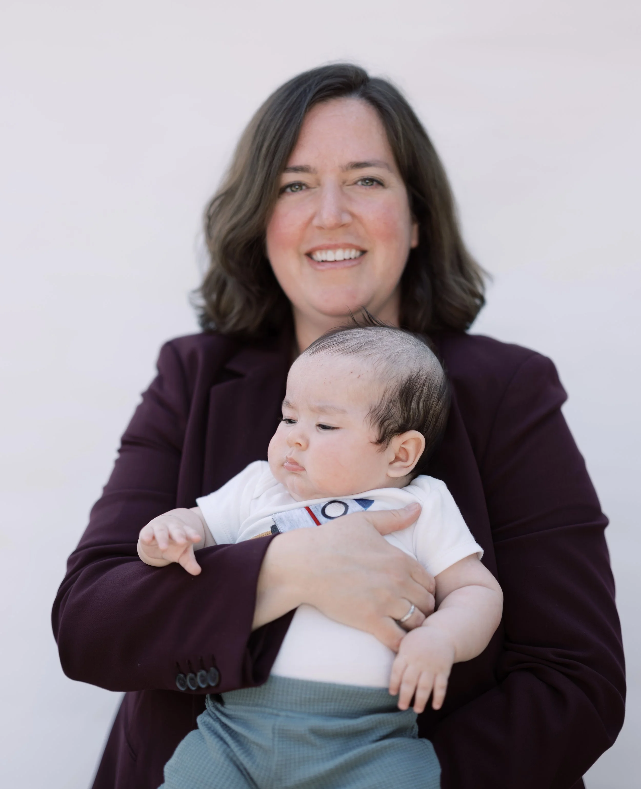 A woman with shoulder-length brown hair, smiling and winking at the camera, holding a baby with black hair and light skin, whose face is turned slightly downward.