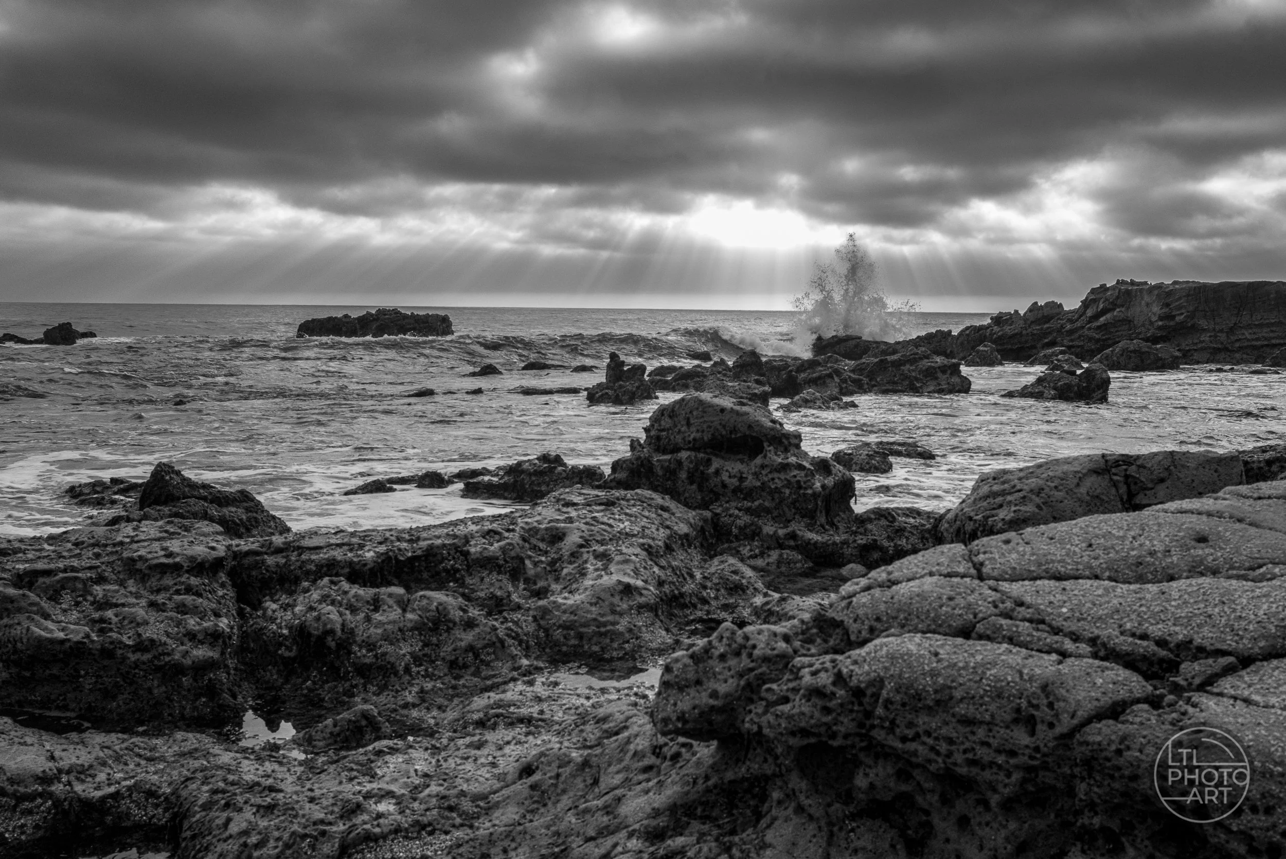 View Black-and-white photograph of sunrays breaking through clouds over the Pacific at Monument Point in Laguna Beach, with a wave splash near the upper-right third of the frame.