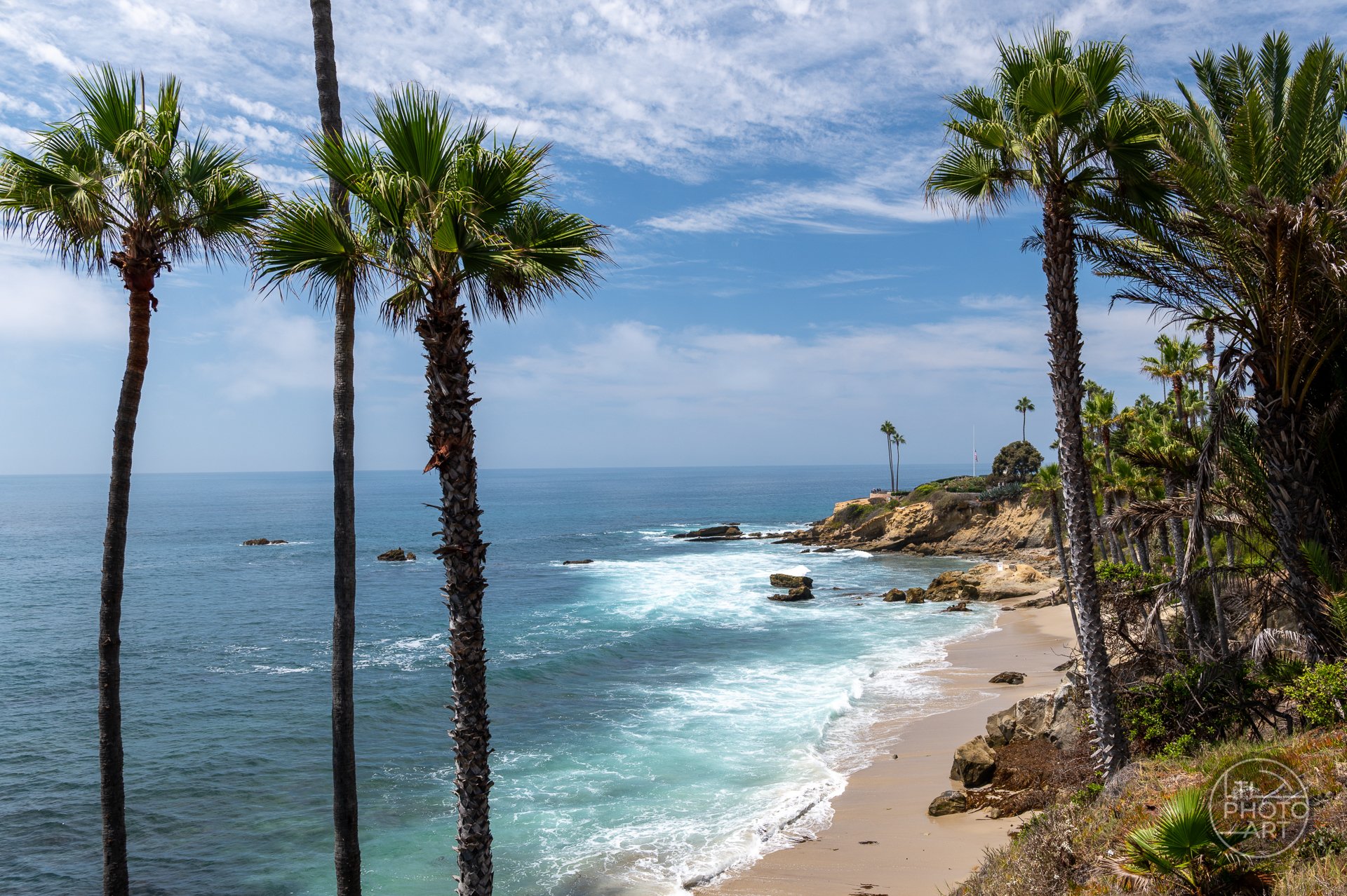 View from the Heisler Park walkway looking north over Rockpile Beach toward Monument Point, framed by palm trees in Laguna Beach, California.