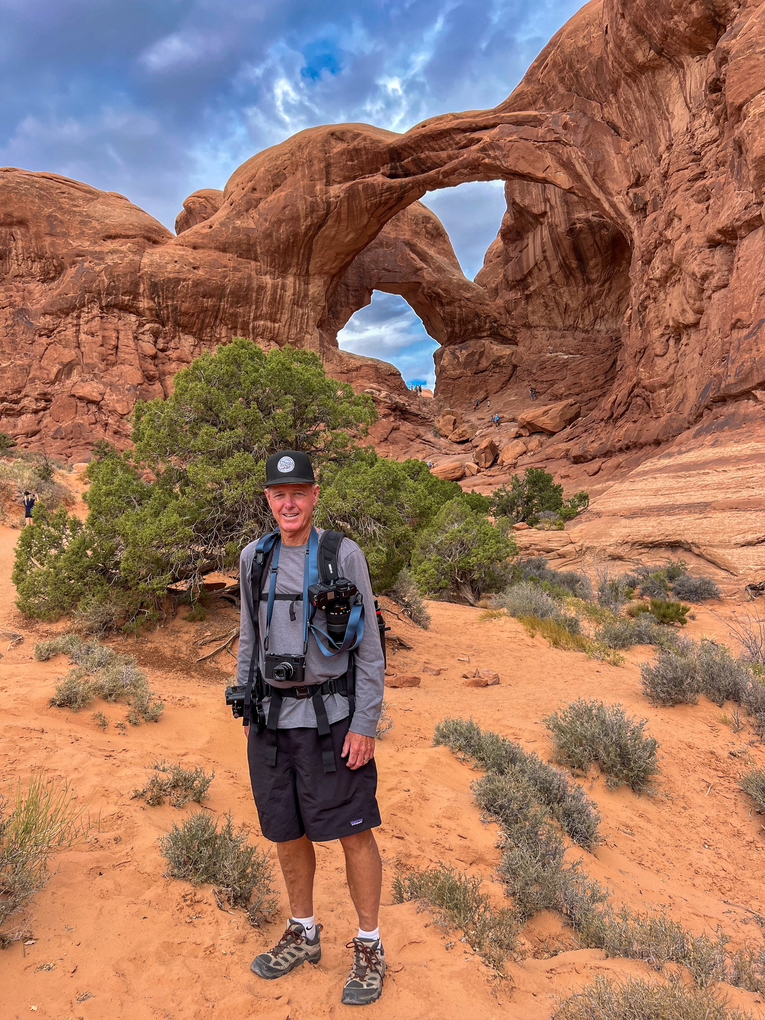 Lyle Lansdell, founder of LTL Photo Art, standing beneath Double Arch in Arches National Park, Utah, wearing multiple cameras while photographing the American Southwest.